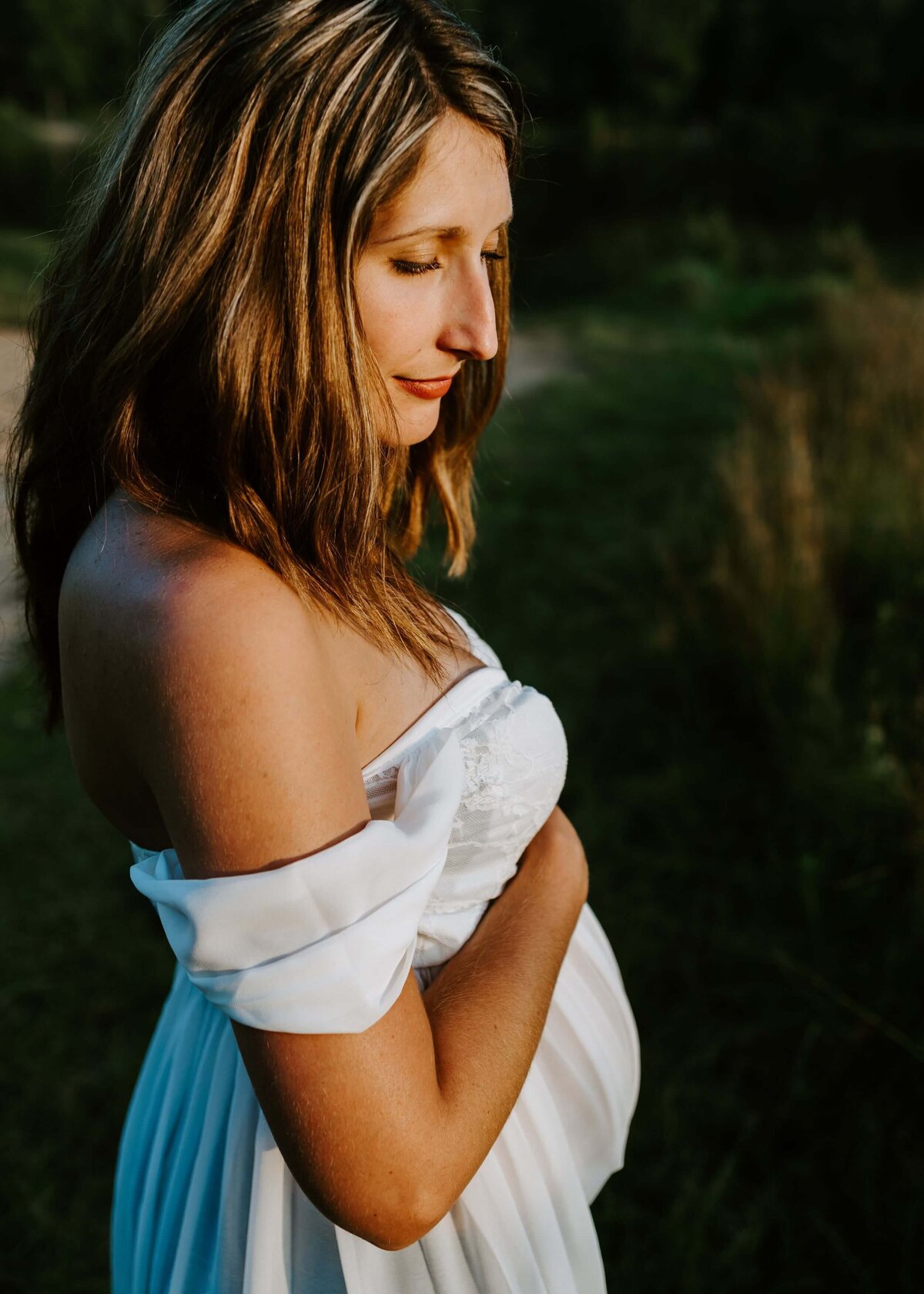 A pregnant woman in a white dress poses elegantly in a field, captured by a Pittsburgh maternity photographer.