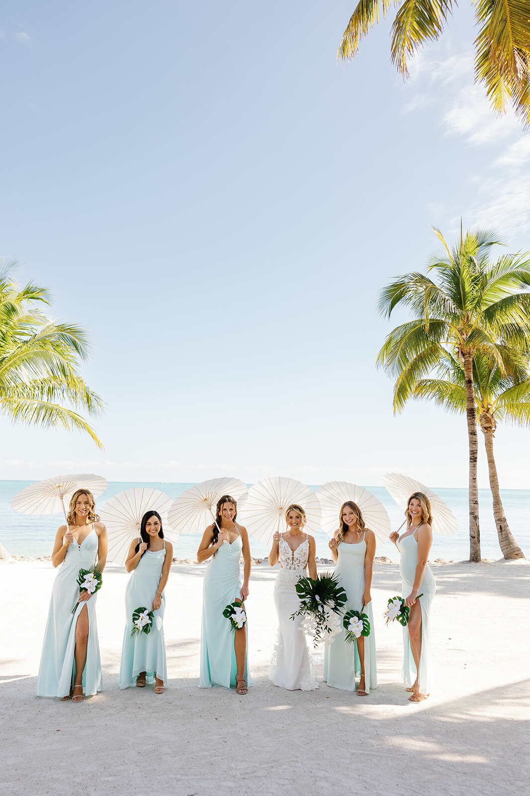 Ashley’s bridesmaids in pastel blue dresses stand in a row holding tropical bouquets with the ocean in the background.