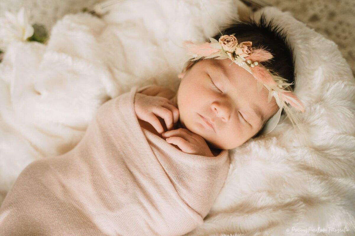 Baby with floral headband – Newborn baby wrapped in a pale blush swaddle, wearing a delicate dried-flower headband, sleeping on a plush white fur blanket.