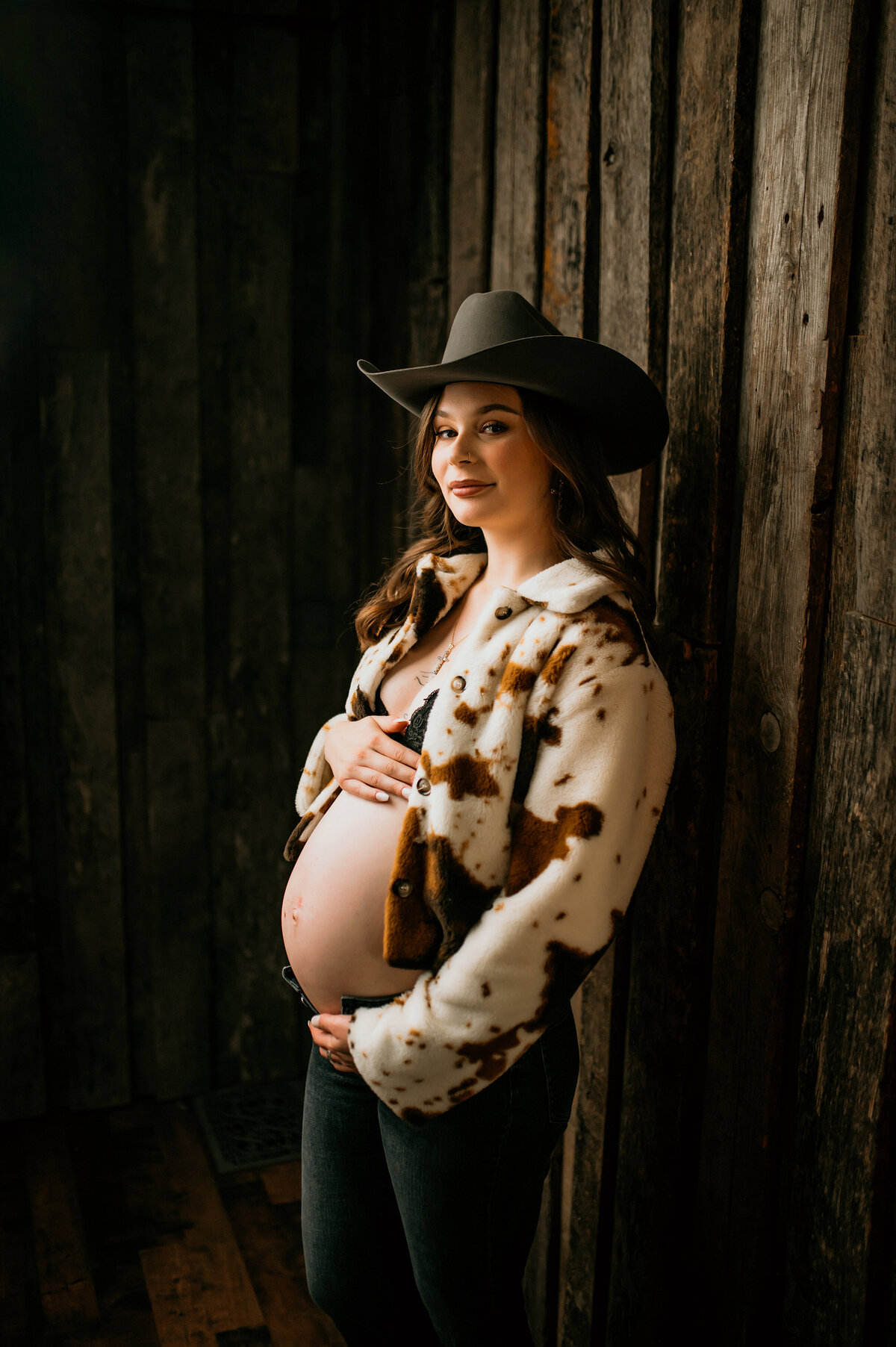 Pregnant woman in a cow-print jacket and cowboy hat, posing confidently against a rustic wooden wall during a western-style maternity session near Oconomowoc, Wisconsin.