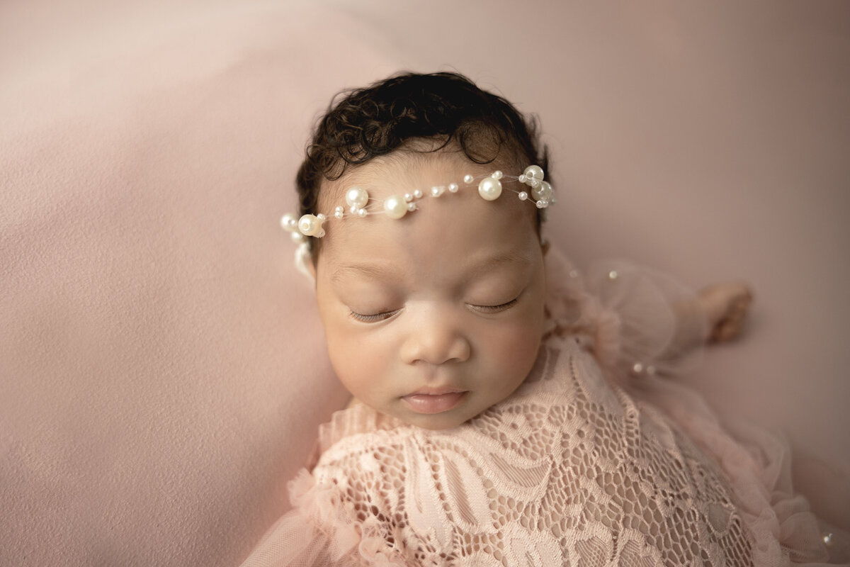 A baby girl sleeps on her back wearing a pearl headband and a pink lace outfit.