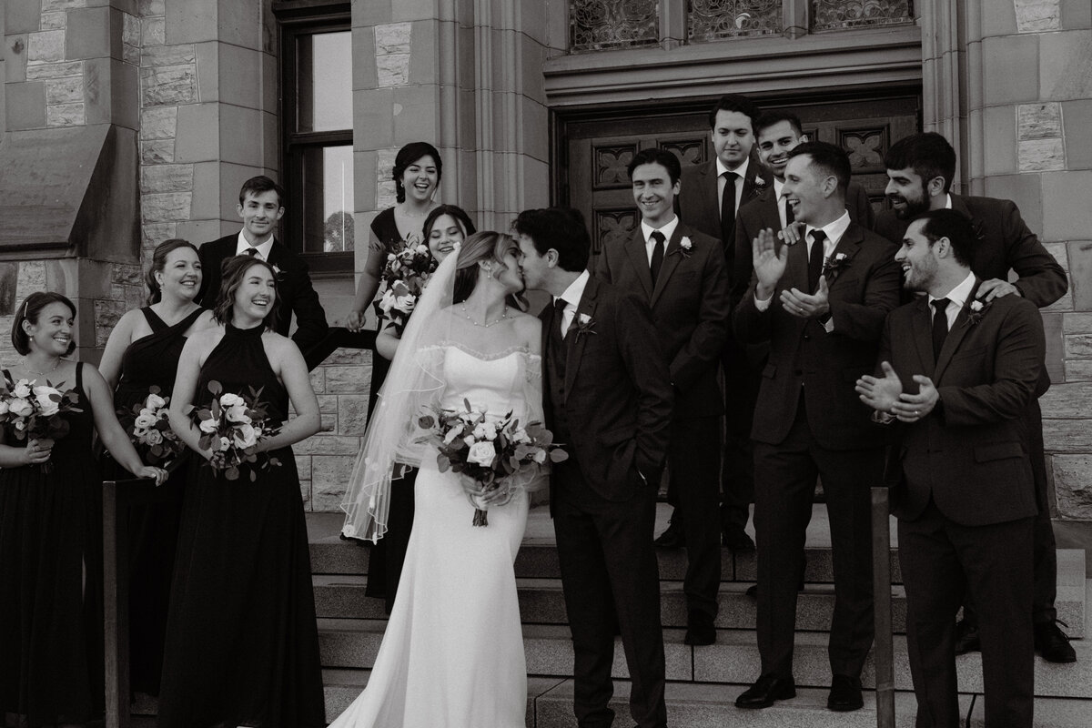 Lovers share a kiss surounded by their wedding party cheering them on