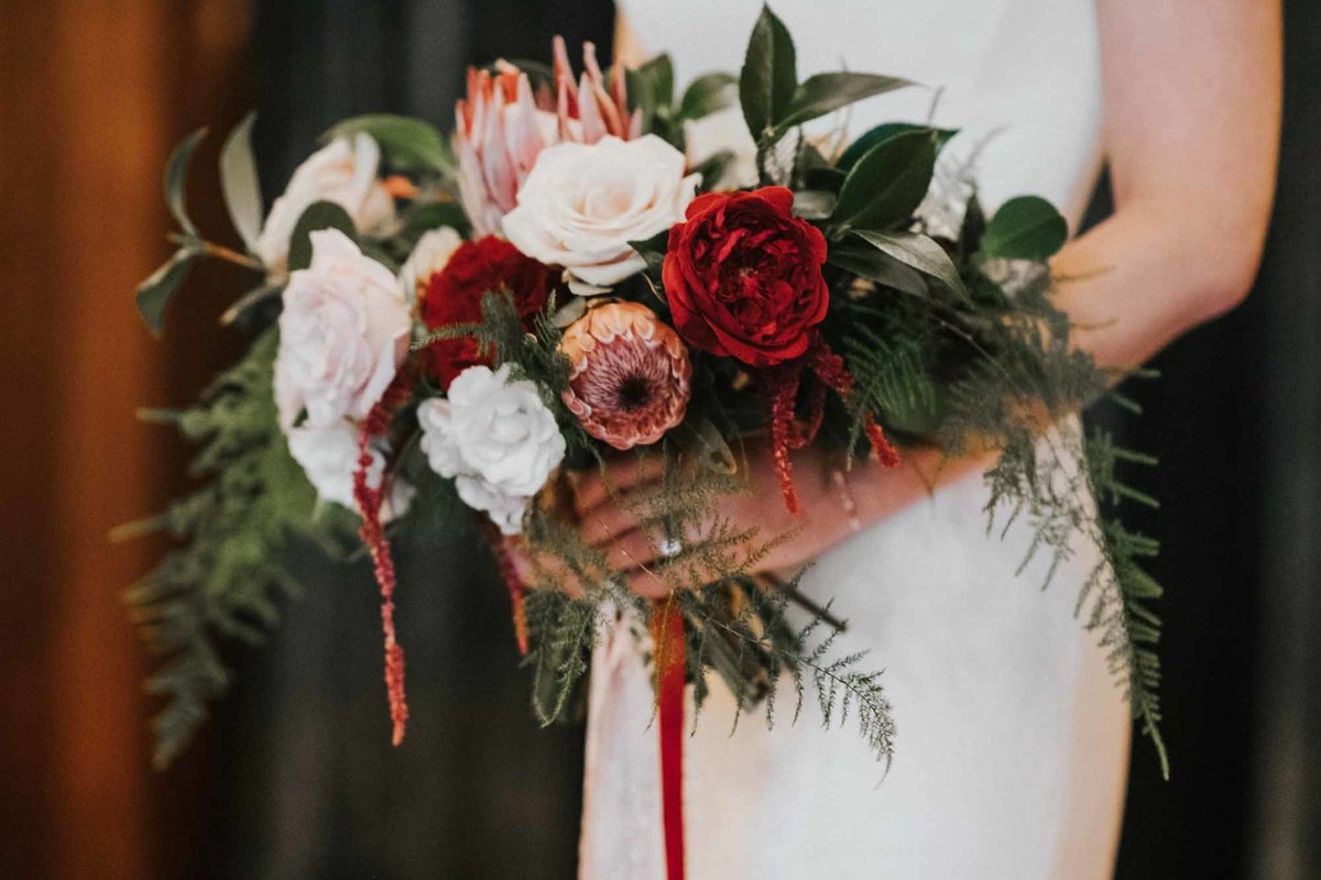 bridal bouquet of red garden roses, pink proteas, blush roses, greenery and long ribbon streamers and trailing greenery