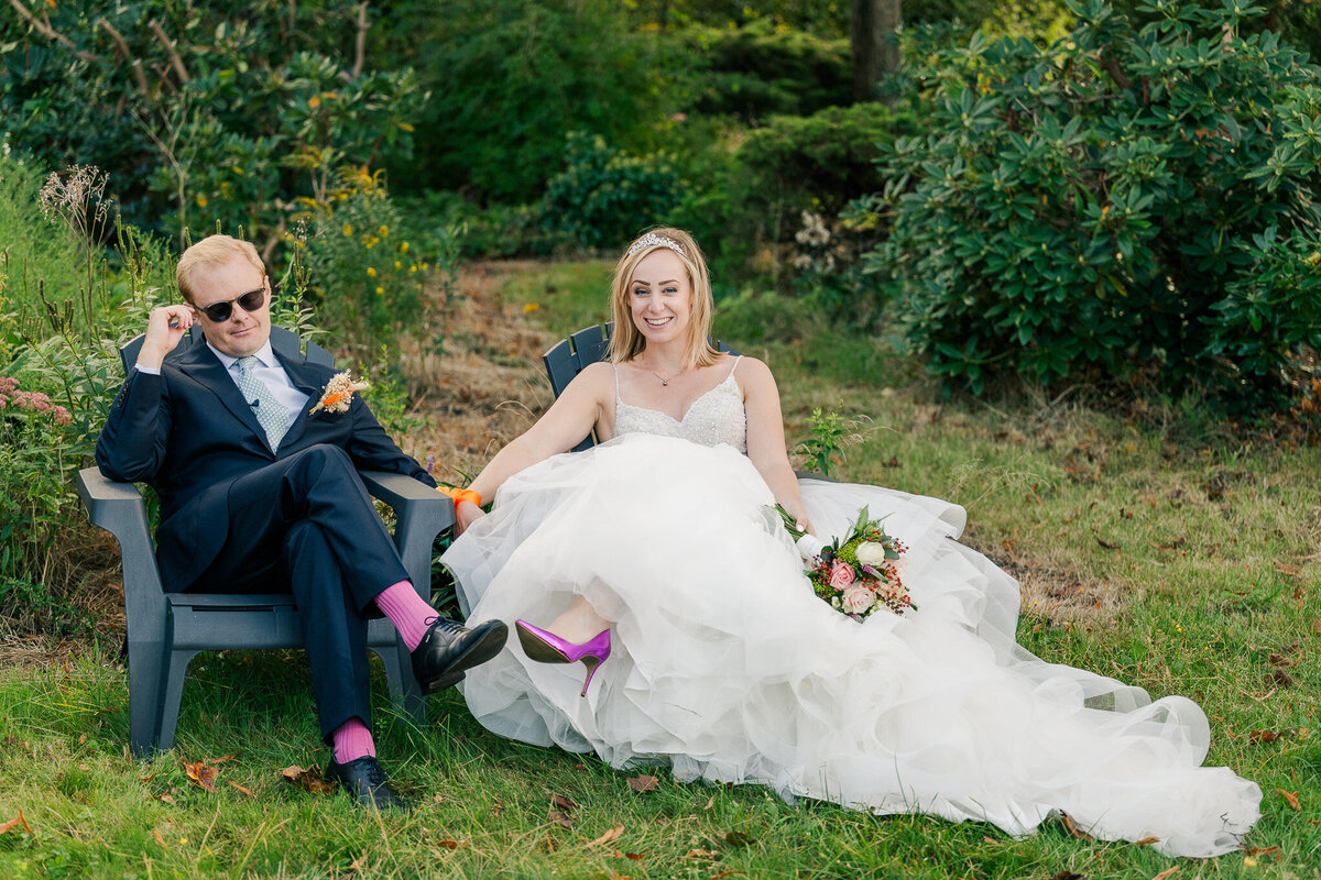 A bride and groom sit in lawn chairs to relax after their garden wedding ceremony. The groom wears sunglasses and his pink socks match the bride's pink shoes