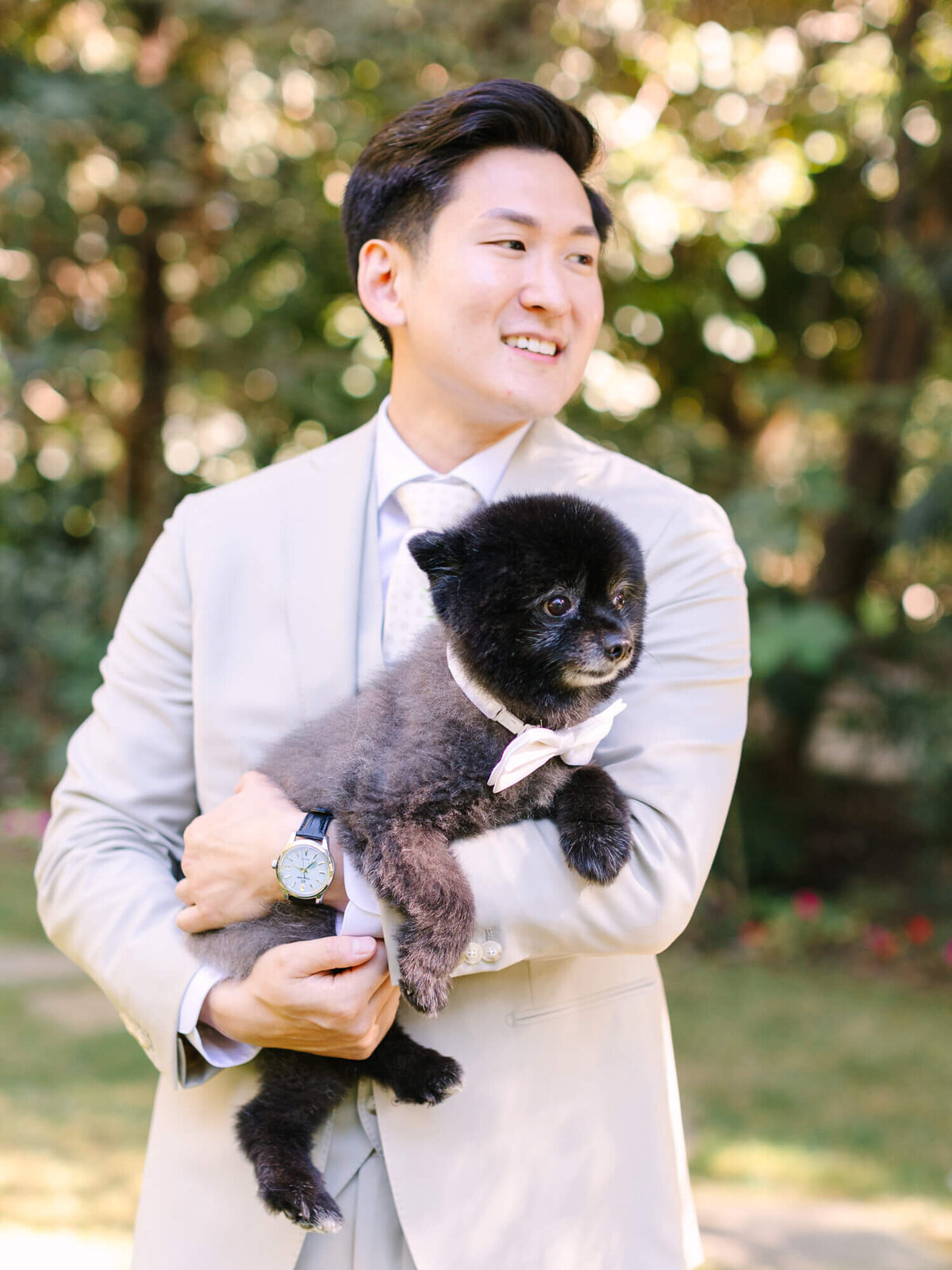 A groom in a light beige suit holds a fluffy black dog wearing a bow tie. They are outdoors with lush greenery.