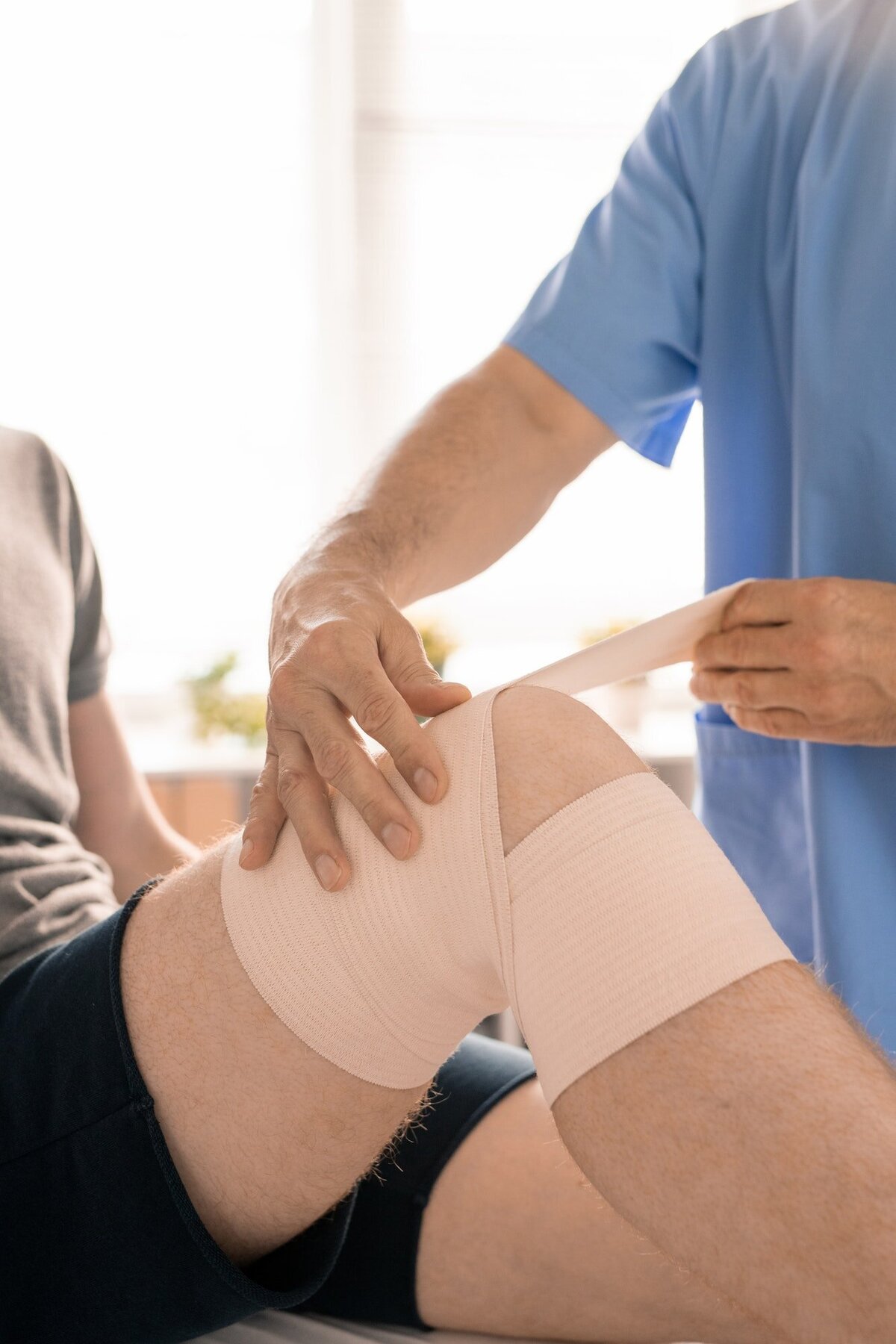 hands-of-clinician-in-blue-uniform-wrapping-injured-knee-of-young-patient