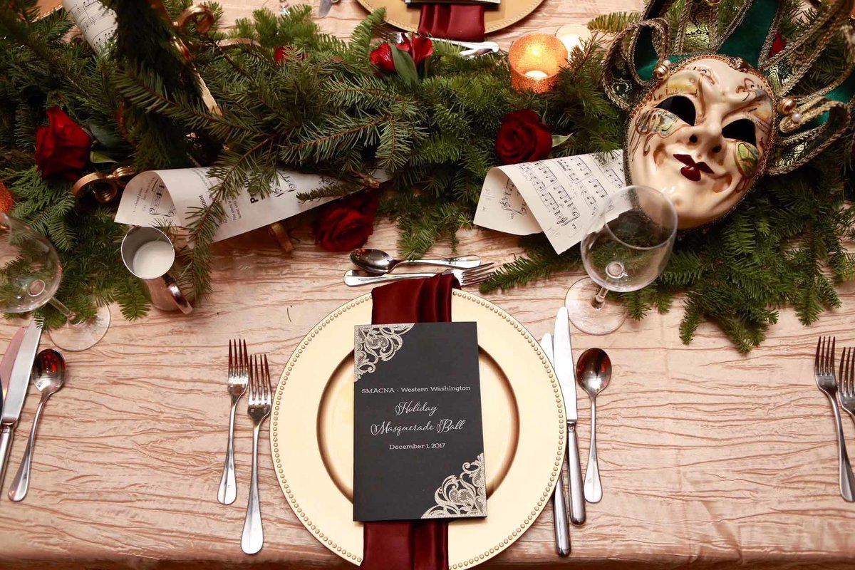 place setting with gold charger plate, red napkin and centerpiece of evergreen garland, red roses, carnival masks on gold linen for holiday party
