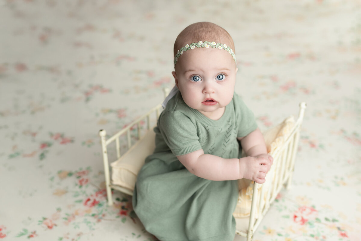 Vintage-inspired milestone of baby girl in sage dress sitting in a floral mini bed—styled softly with pastel tones.