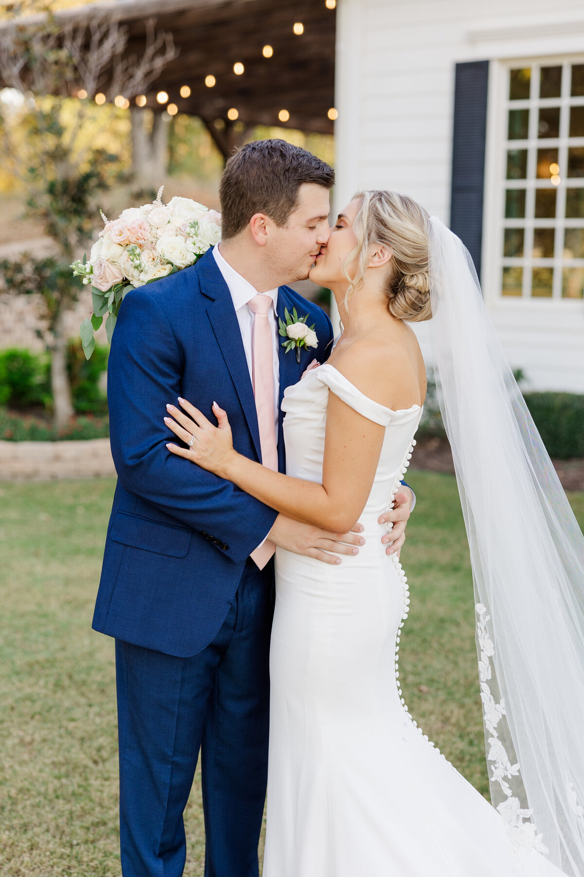 A bride and groom share a kiss outside a white chapel adorned with string lights at a romantic outdoor wedding near McKinney, Texas. The bride’s fitted gown and veil complement the groom’s navy suit and blush tie.