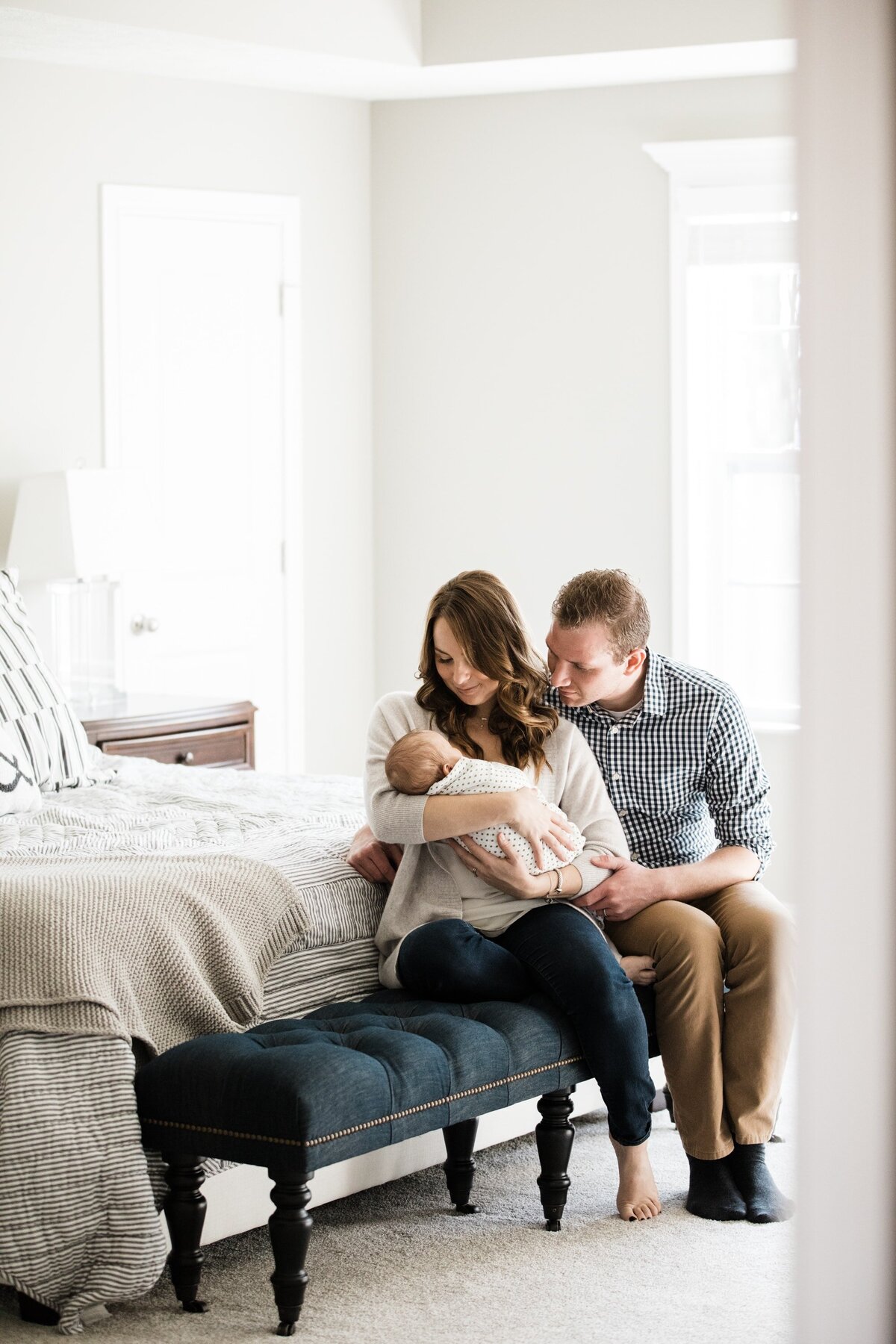 A couple sitting closely on a bedroom bench, affectionately looking at their baby, captured perfectly by their Pittsburgh newborn photographer.