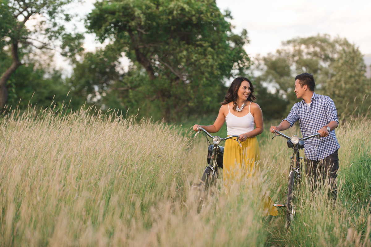 engaged couple walking their vintage bikes through the tall grass at sunset
