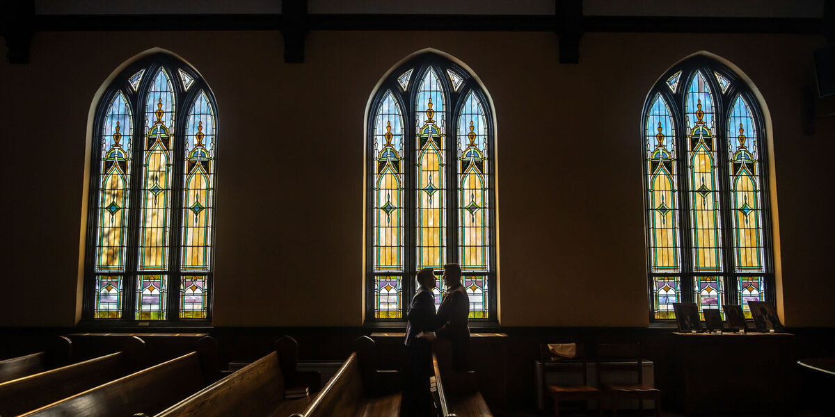Groom and groom are standing in front of a stained glass window in a church.