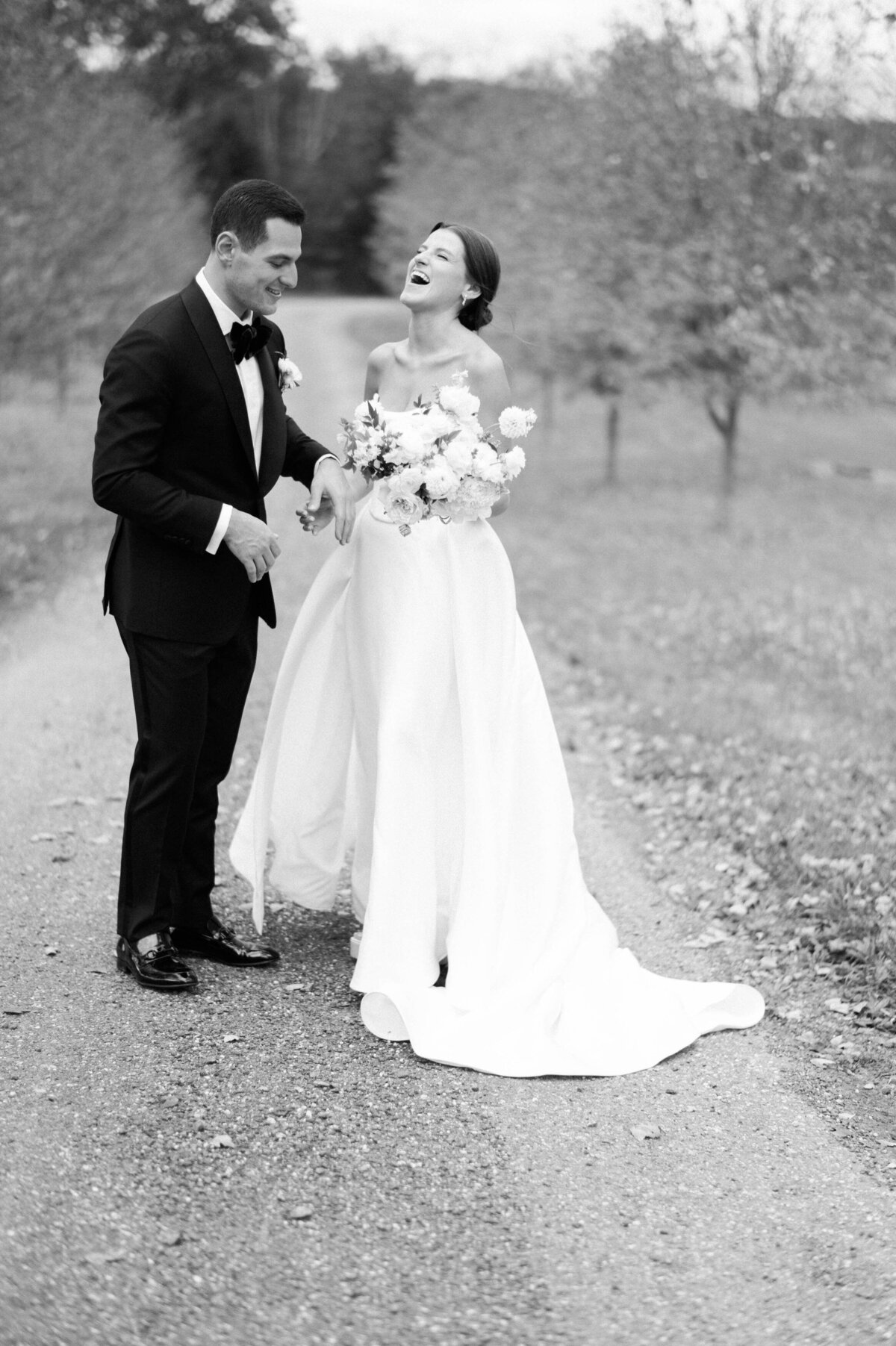 the bride and groom laughing together on a gravel path, surrounded by trees