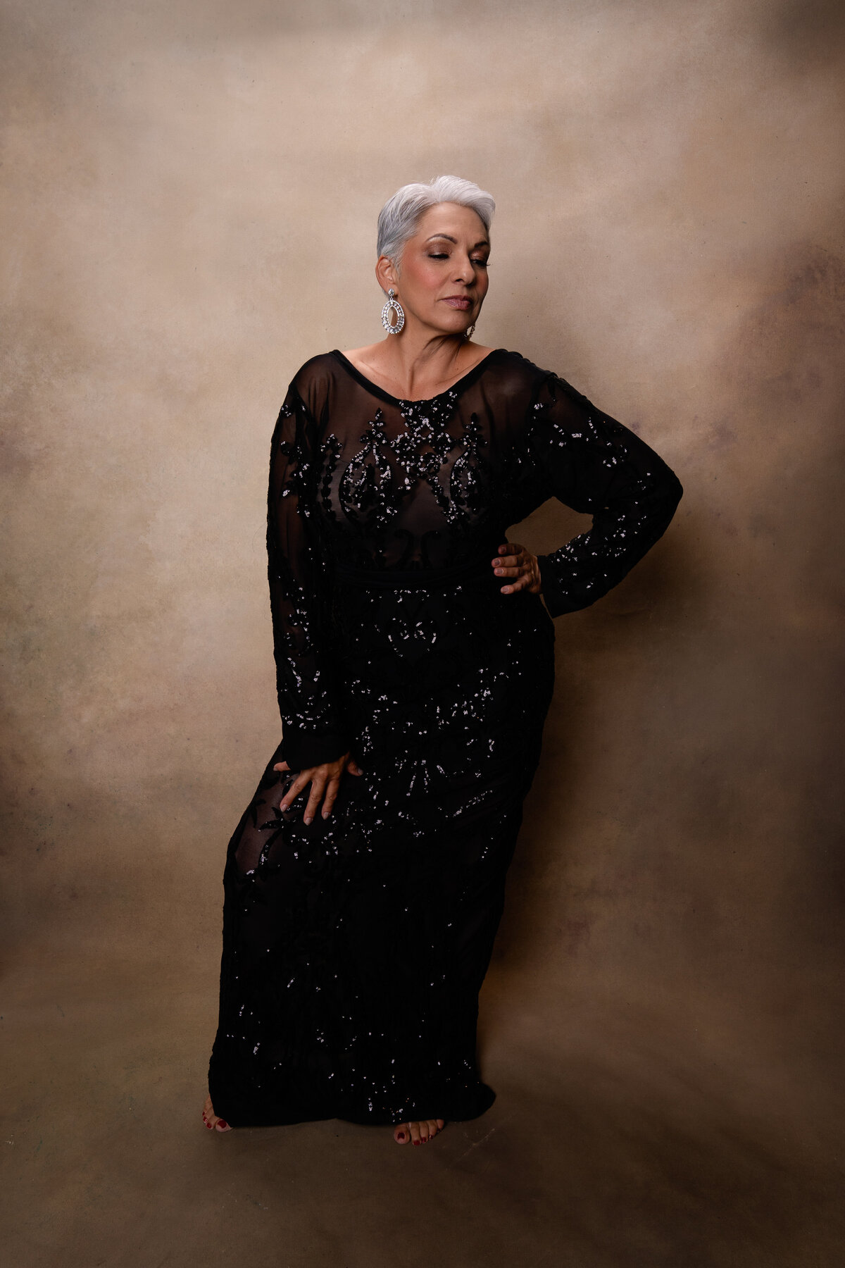 Middle Aged woman with short gray hair standing in black dress on a beige backdrop.
