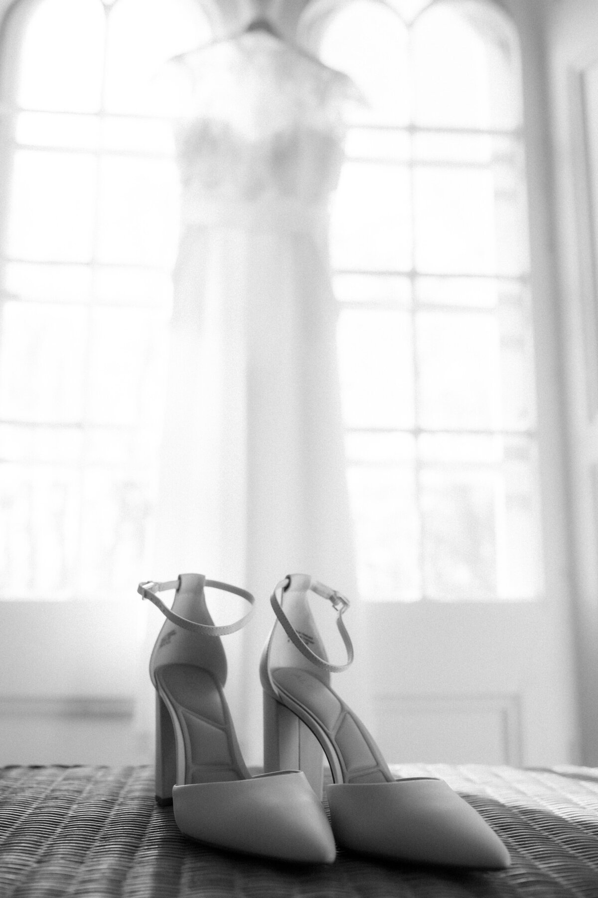 black and white image of a bride's shoes with her dress hanging in a window behind