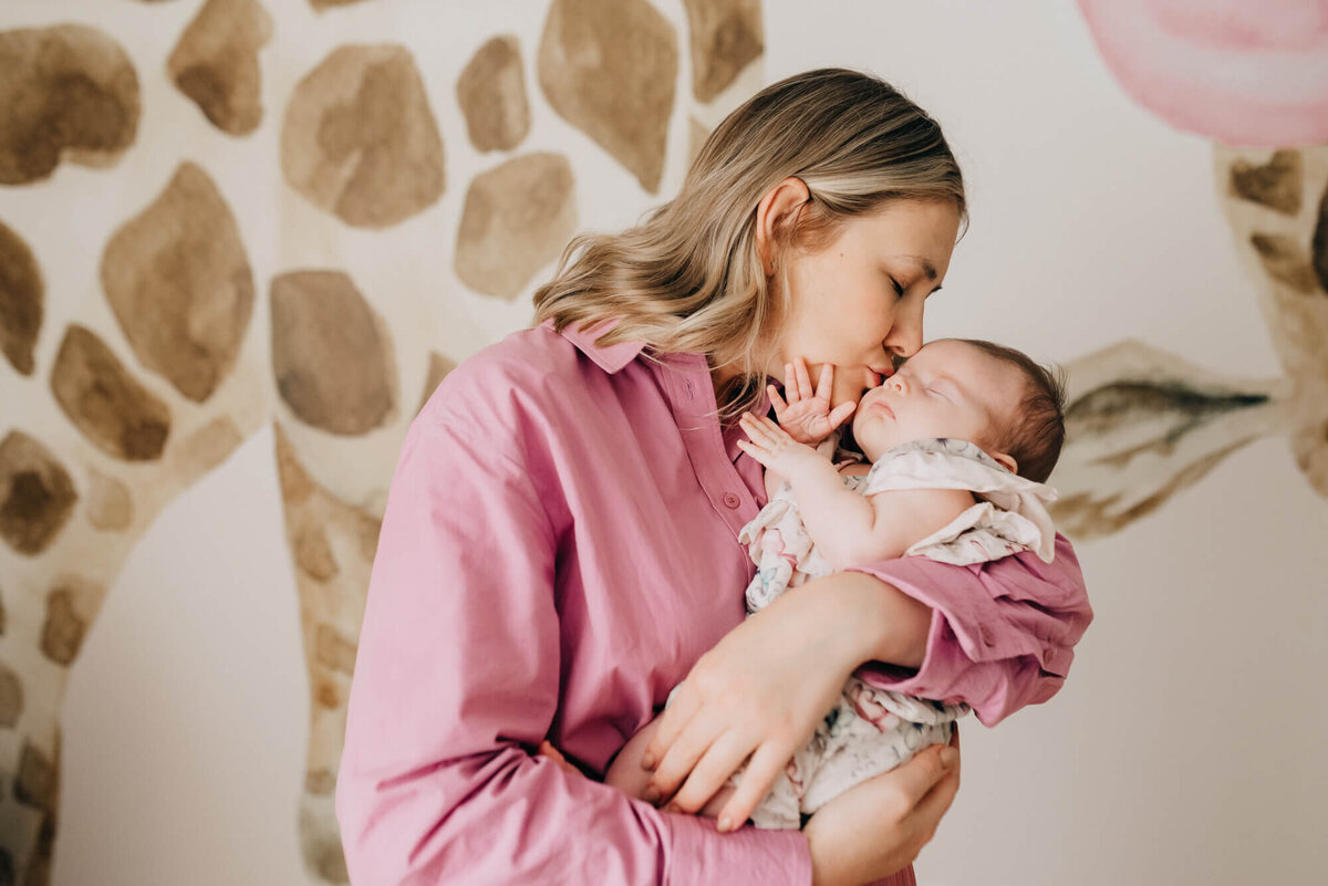 A mother wearing a soft pink shirt kisses her newborn daughter in her safari themed nursery
