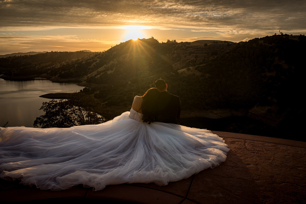 A breathtaking wedding portrait of a bride and groom embracing on the edge of a cliff at Airola Road Vineyard, overlooking the Sierra Nevada foothills and New Melones Reservoir during a stunning golden sunset.