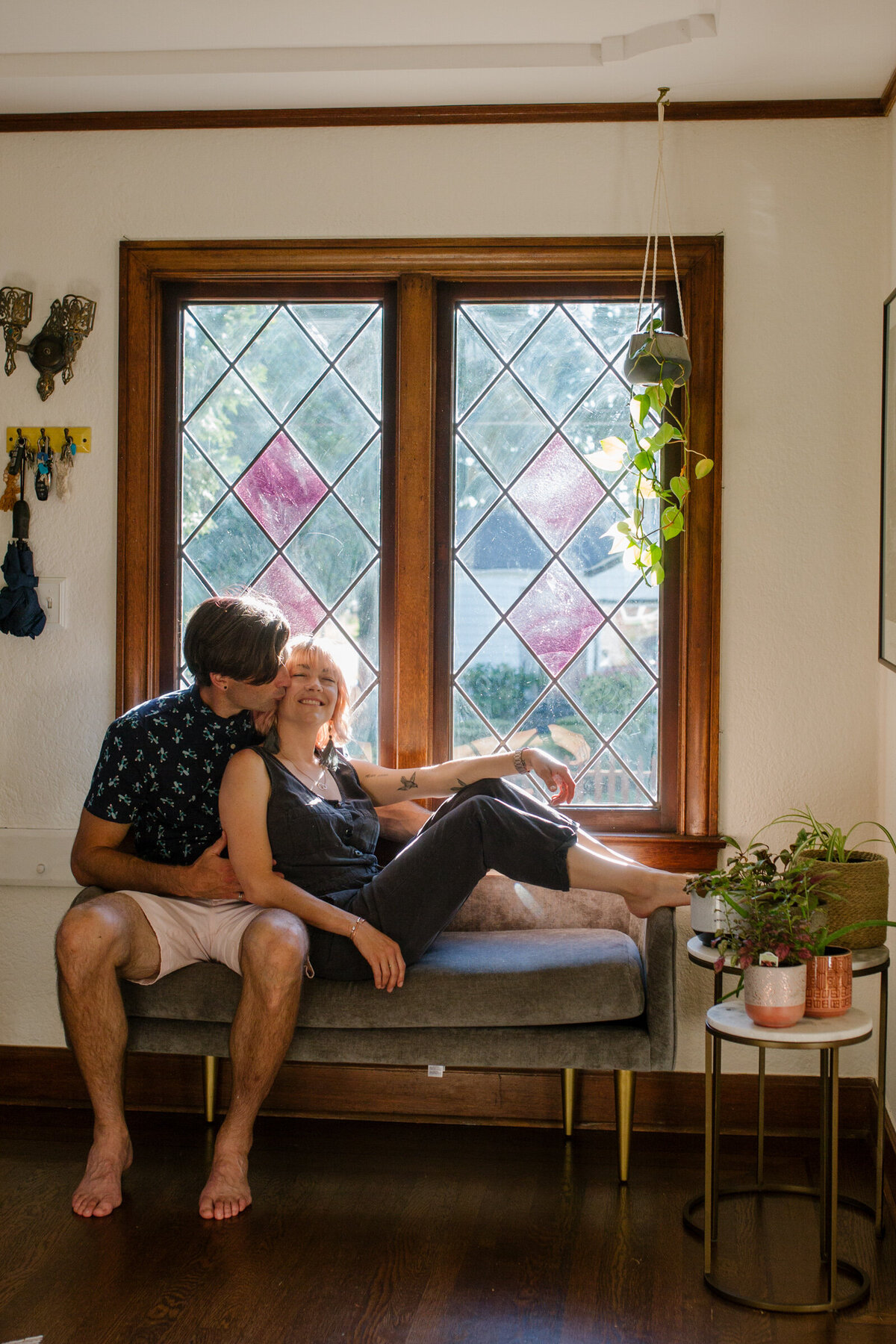 A couple cuddling on a couch near a window in their house.