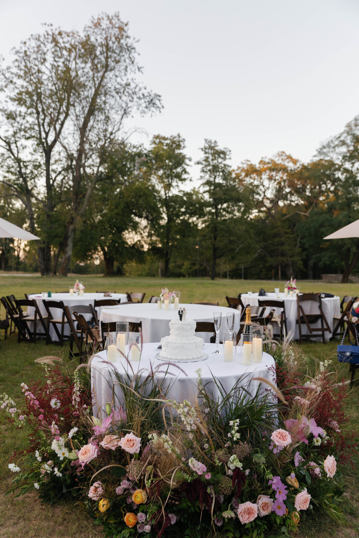 Outdoor wedding reception table decorated with a lush garden-style floral meadow featuring blush roses, pink lilies, ranunculus, astilbe, and textured greenery surrounding a small white vintage wedding cake. Candles and champagne sit on the table with round guest tables in the background, set in an open-air field surrounded by trees at sunset. Designed by a luxury wedding florist specializing in romantic, organic arrangements.