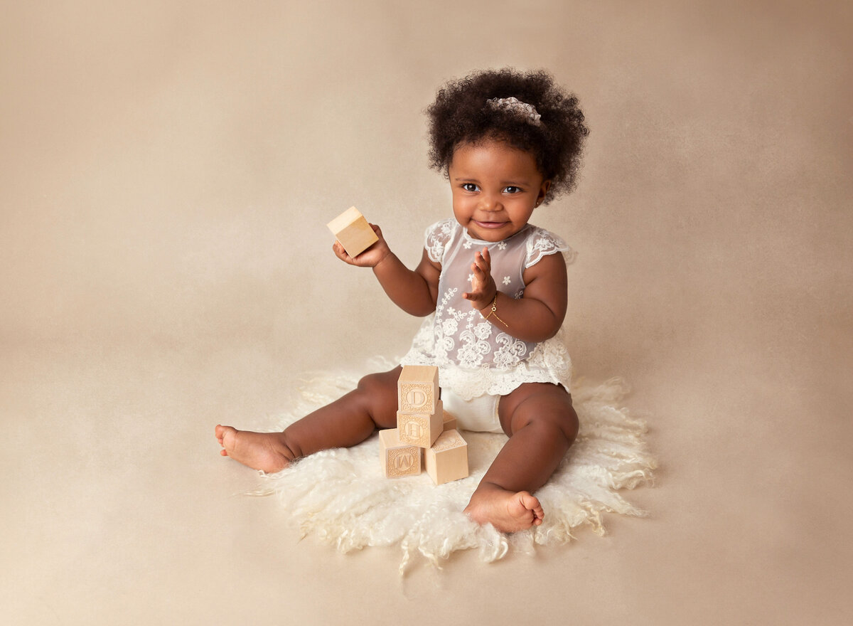 Baby girl in white lace romper is sitting for a 6-month baby milestone photoshoot. She has wooden blocks stacked between her legs and is smiling at the camera.