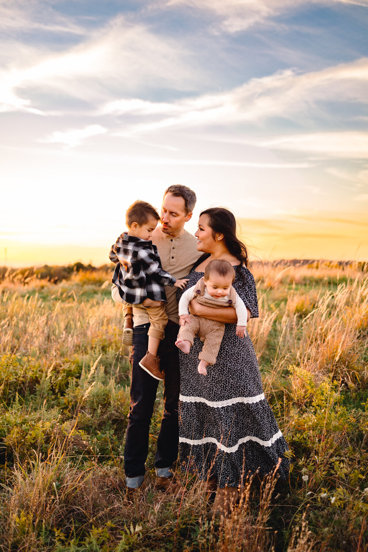 Maternity and family portrait sessions in Albuquerque capture a beautiful moment in a forest setting. The woman, wearing a blue dress with white dots, holds a baby while looking at the man, who is dressed in black with coffee-colored accents and is carrying a larger child.