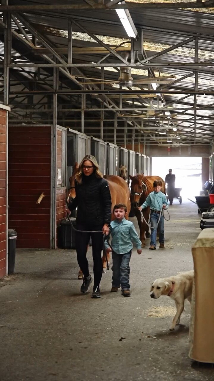 two young riders in matching western shirts lead chestnut ponies through a barn aisle while their riding instructor helps them 