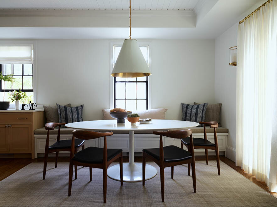 Discover the casual dining room in a historic Hingham home, featuring a custom bench under the window, Saarinen-inspired dining table, mid-century furniture, cone light fixture, and white linen shades and drapery.