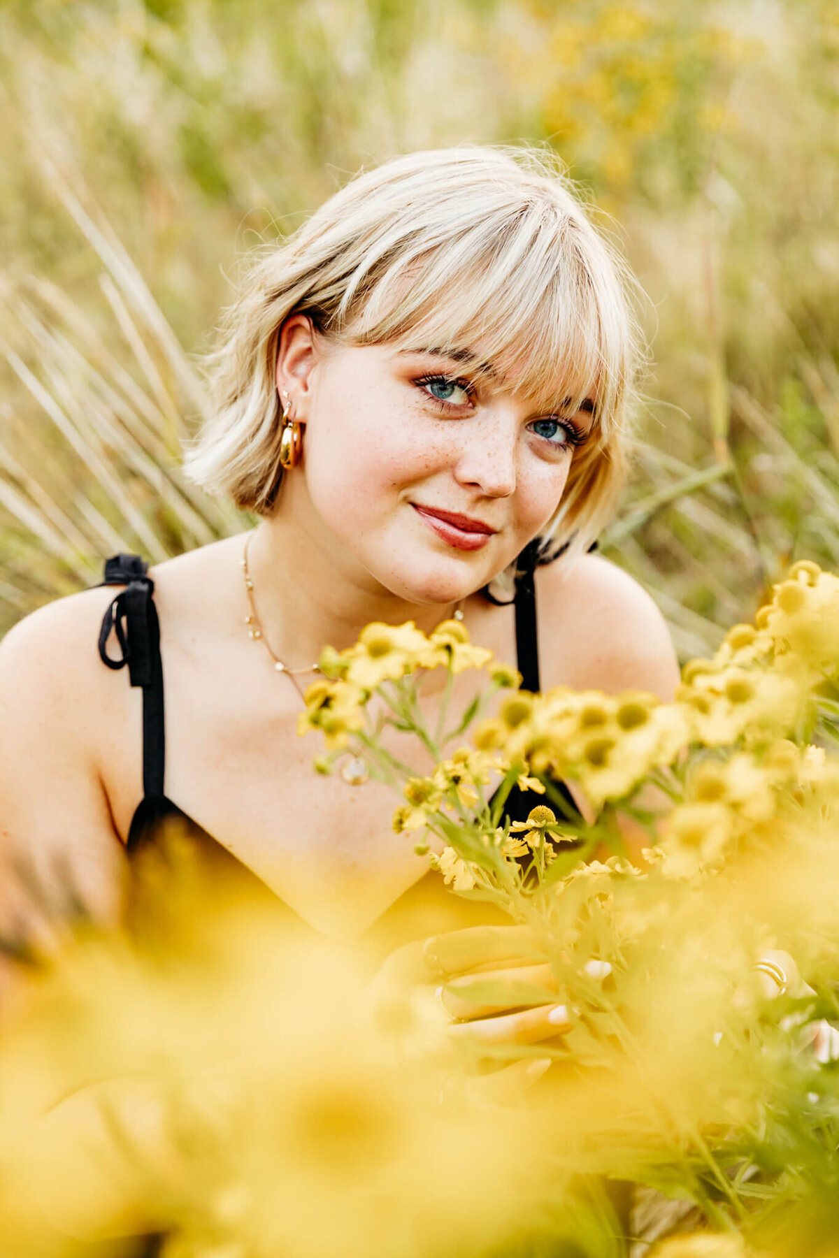 high school girl in a black dress kneeling behind yellow flowers and glancing up with a slight smile captured by Ashley Kalbus Photography