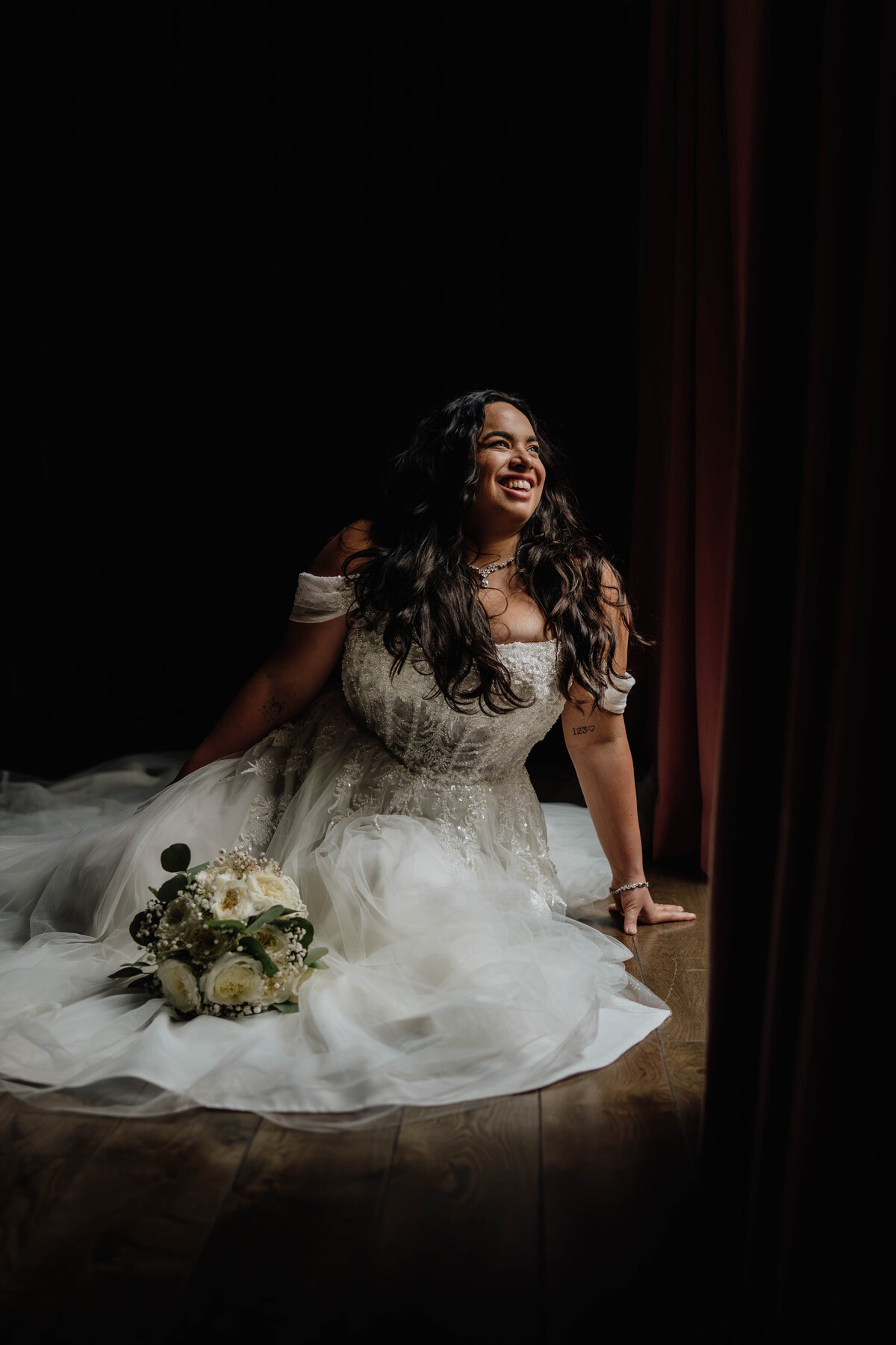 A bride sits happily on the floor in front of a large window. The light is and the background is dark and moody.