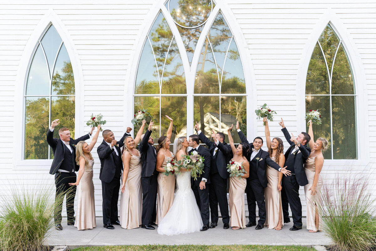 Full wedding party celebrating while bride and groom kiss in front of tall windows at Springs Wallisville
