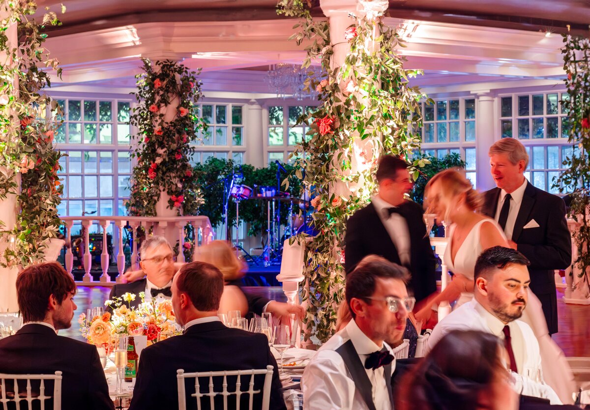 A lively wedding reception in a beautifully decorated venue. Guests in formal attire are seated at tables adorned with floral arrangements, while others stand and converse near columns entwined with greenery and lights.