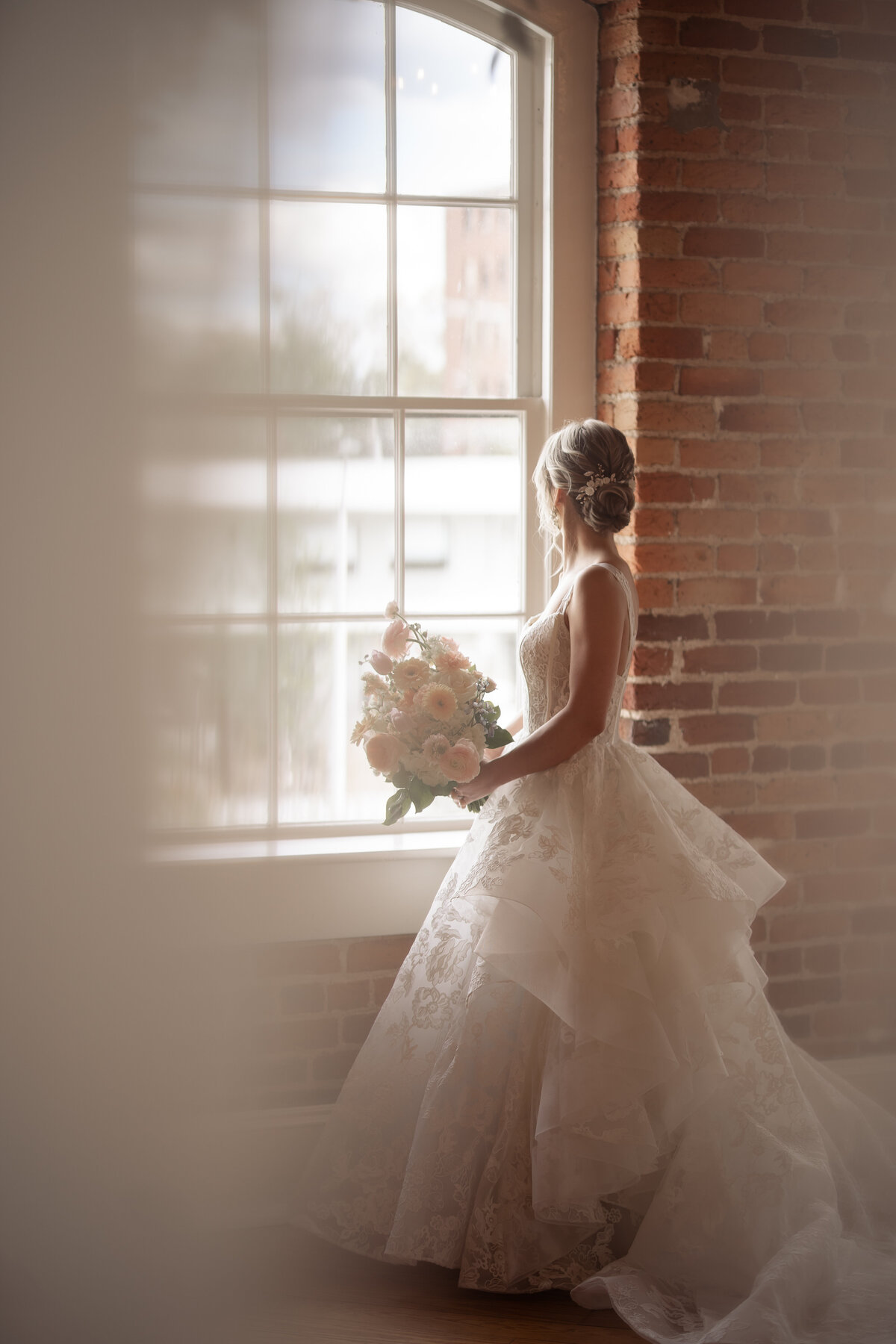 Bride looking out a window with her bouquet and a curtain slightly in the way of the shot