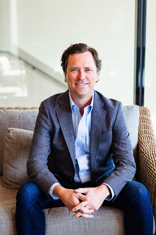 Man in blue jacket leans forward on couch during his headshot session with Angela Marie Media.