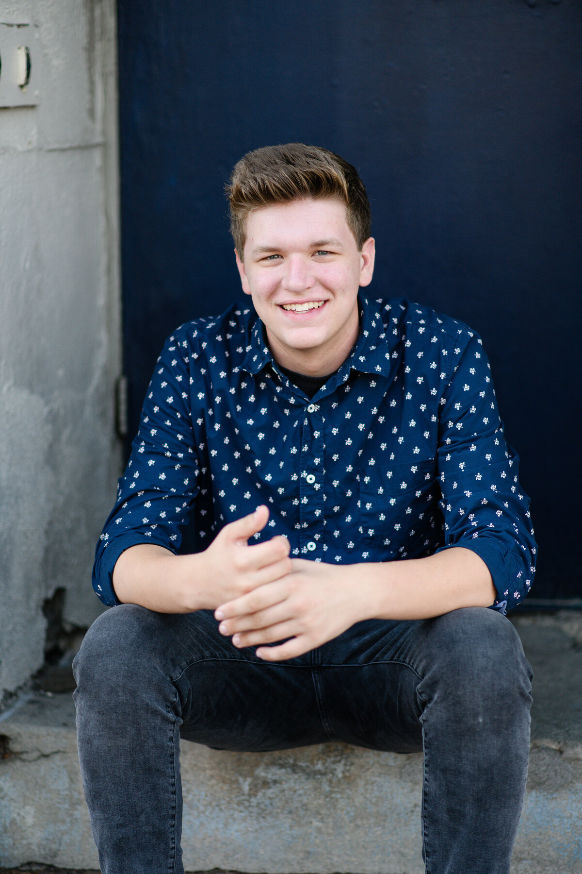 denver senior photographer captures senior guys photo ideas with a young man wearing a dark blue button down while sitting on a stair with her arms resting on his knees  and smiling taken