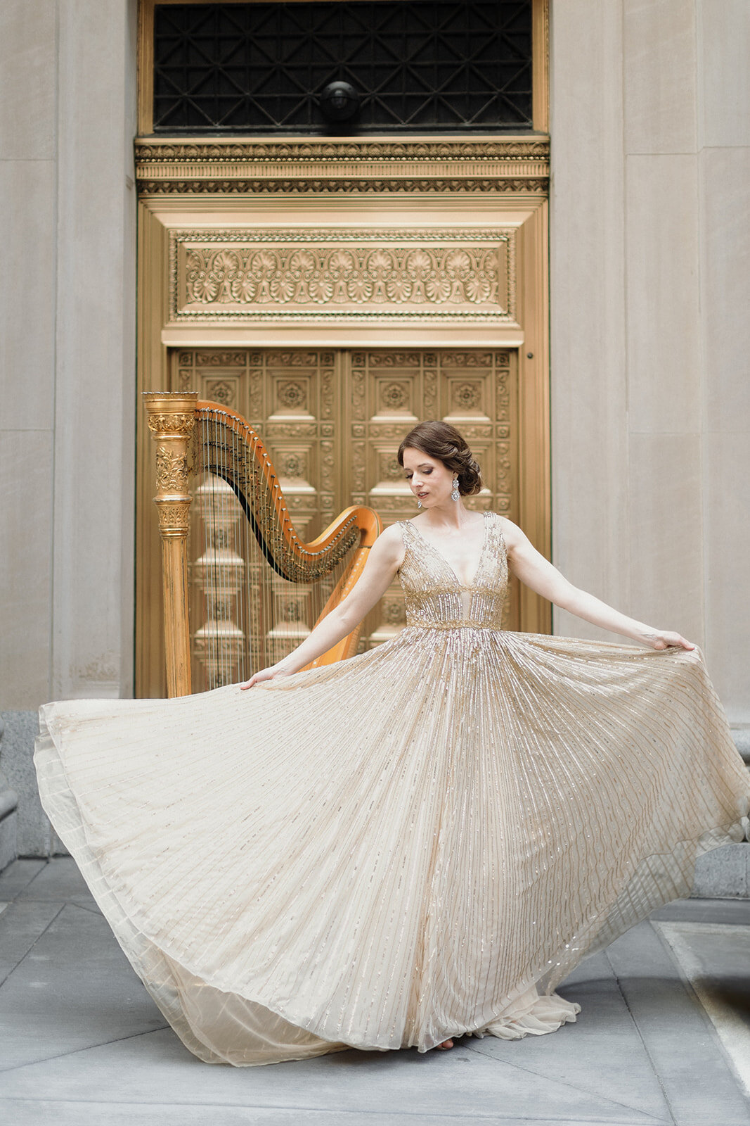 woman in gold gown is fanning out the gown, in front of a gold harp and large gold brass doors in Chicago.