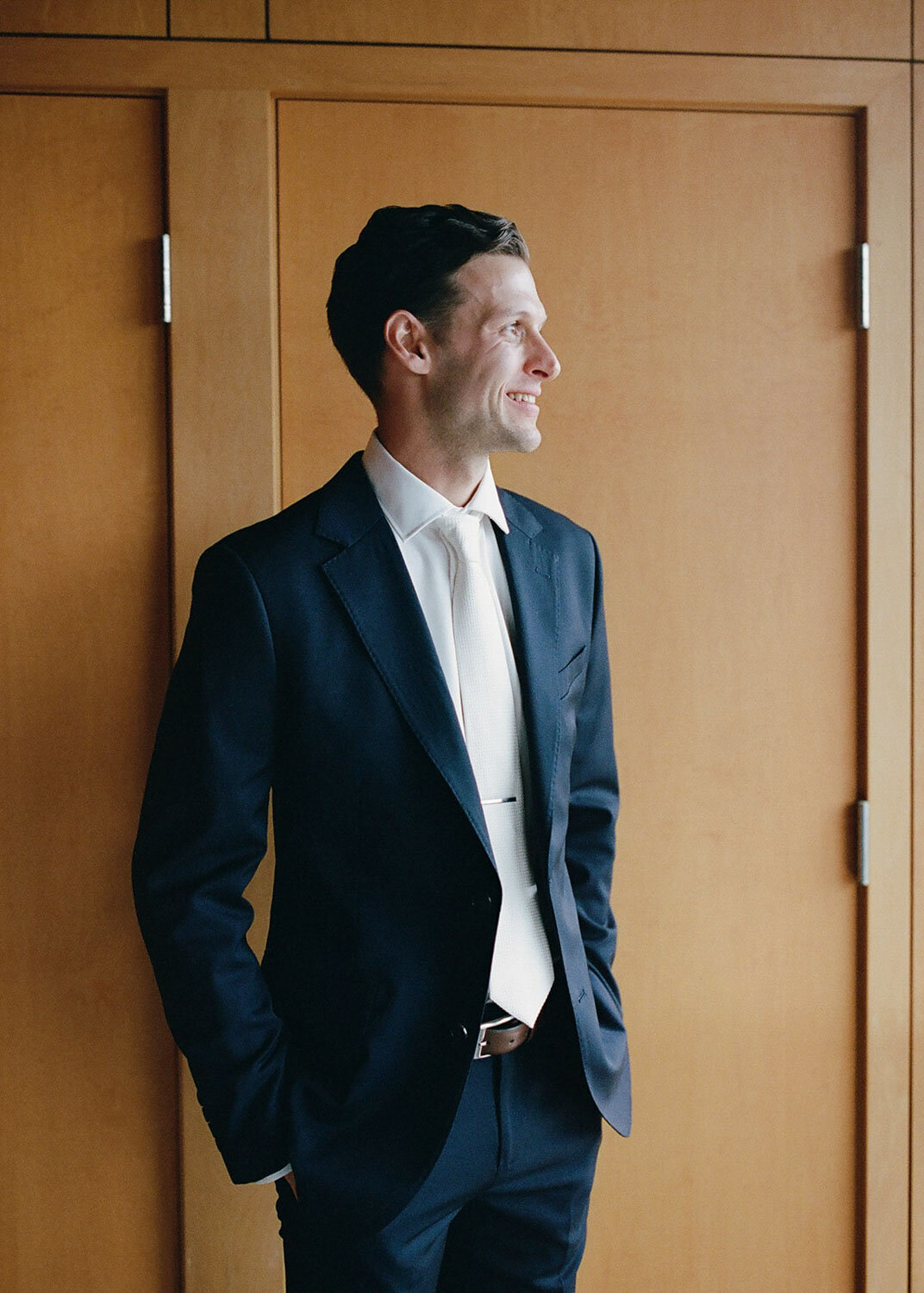 Groom in a navy suit standing by a wooden wall smiling