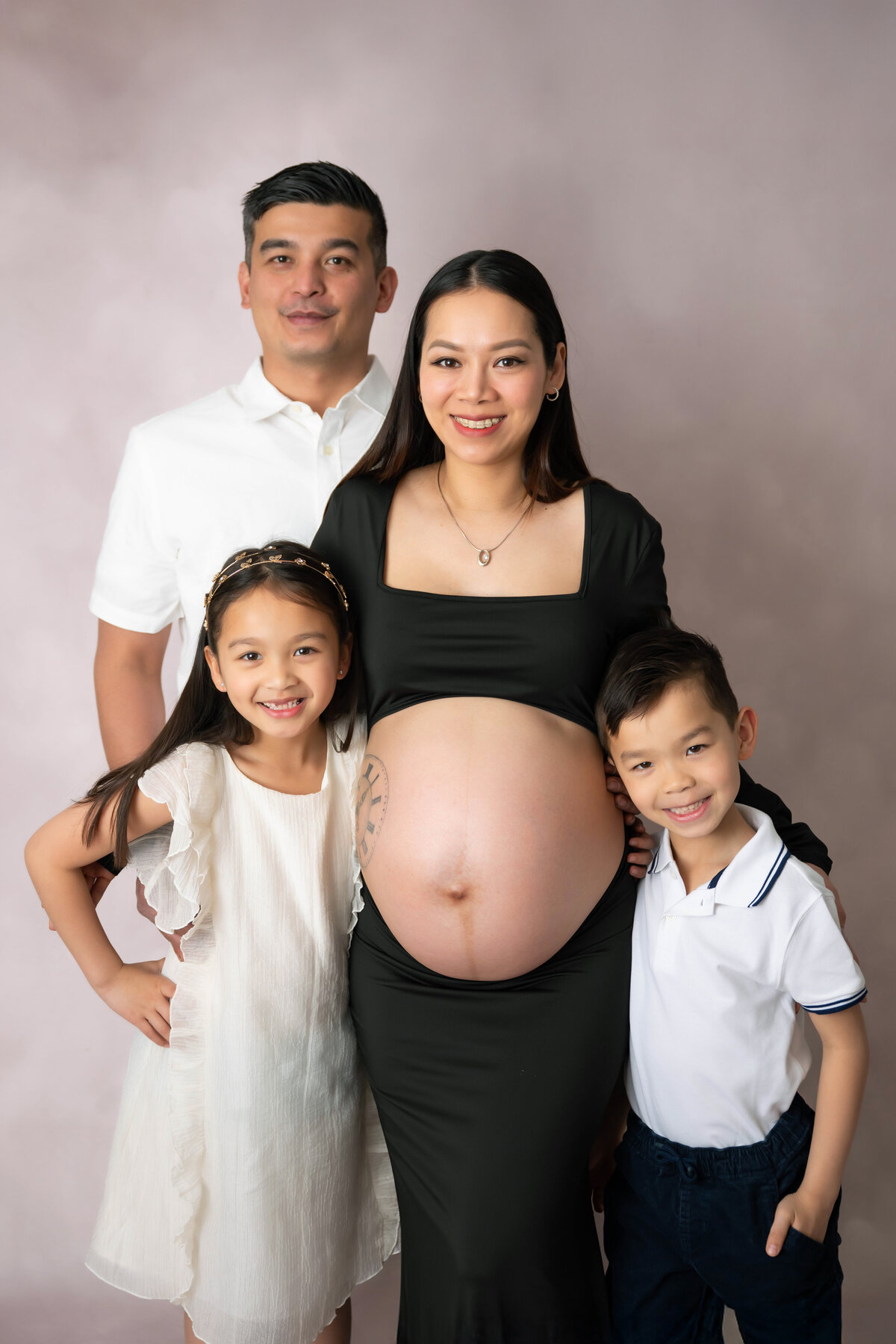 Expecting parents pose as they hug two young children as mom stands with pregnant belly exposed. 