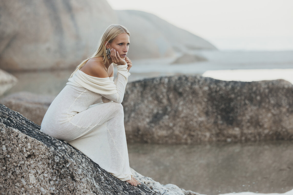Model Johanna zit dromerig op een rots in Kaapstad in een off-shoulder witte jurk, omgeven door zachte zonsopgang en rotslandschap.