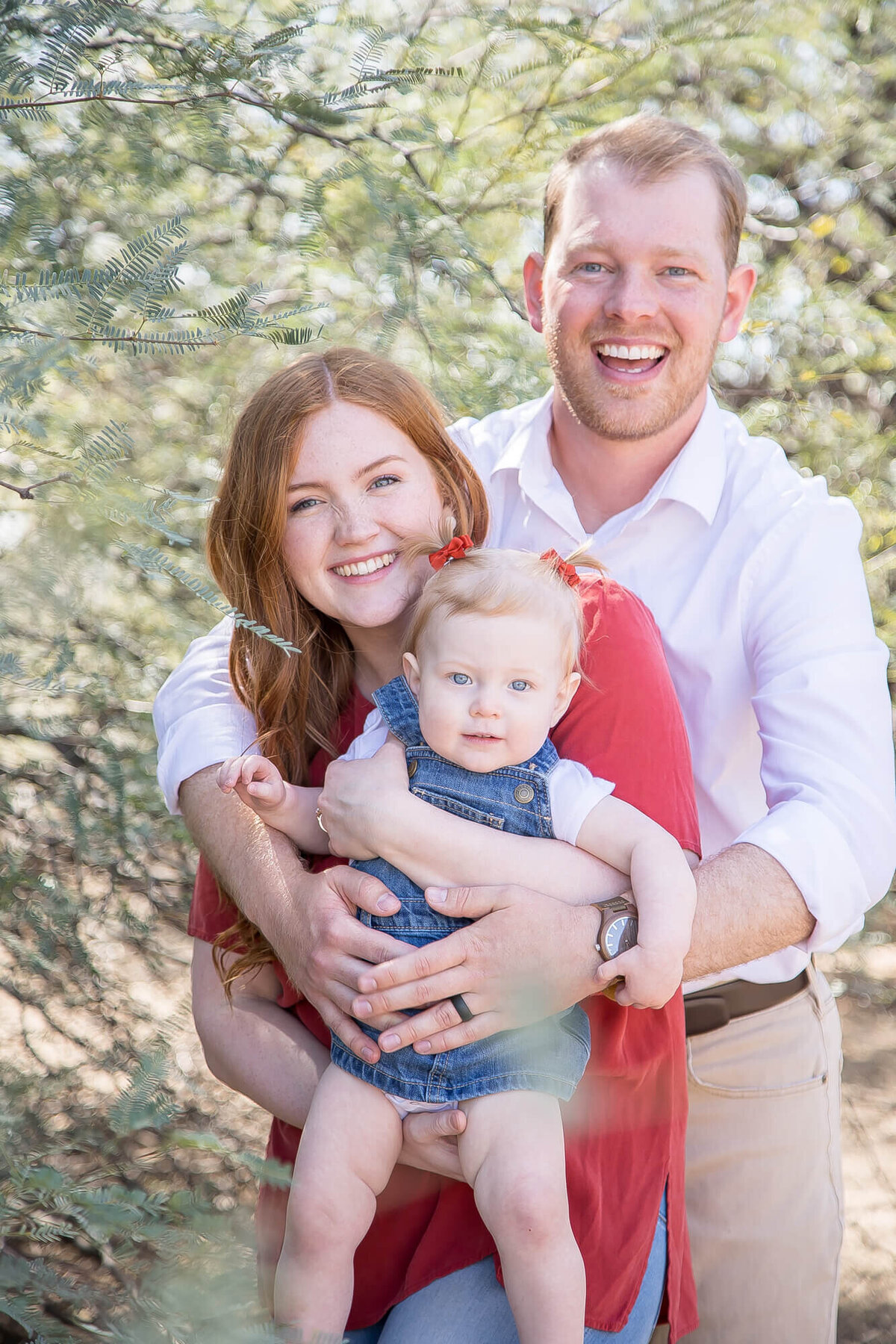 toddler held by red headed mother in a red shirt  and dad in a white shirt