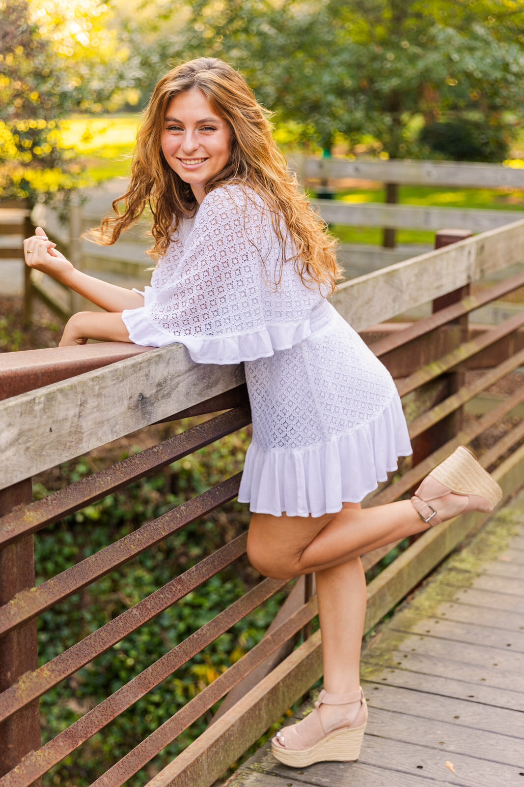 senior leaning on a wooden bridge wearing a white dress one leg popped back up in an Atlanta park during portraits