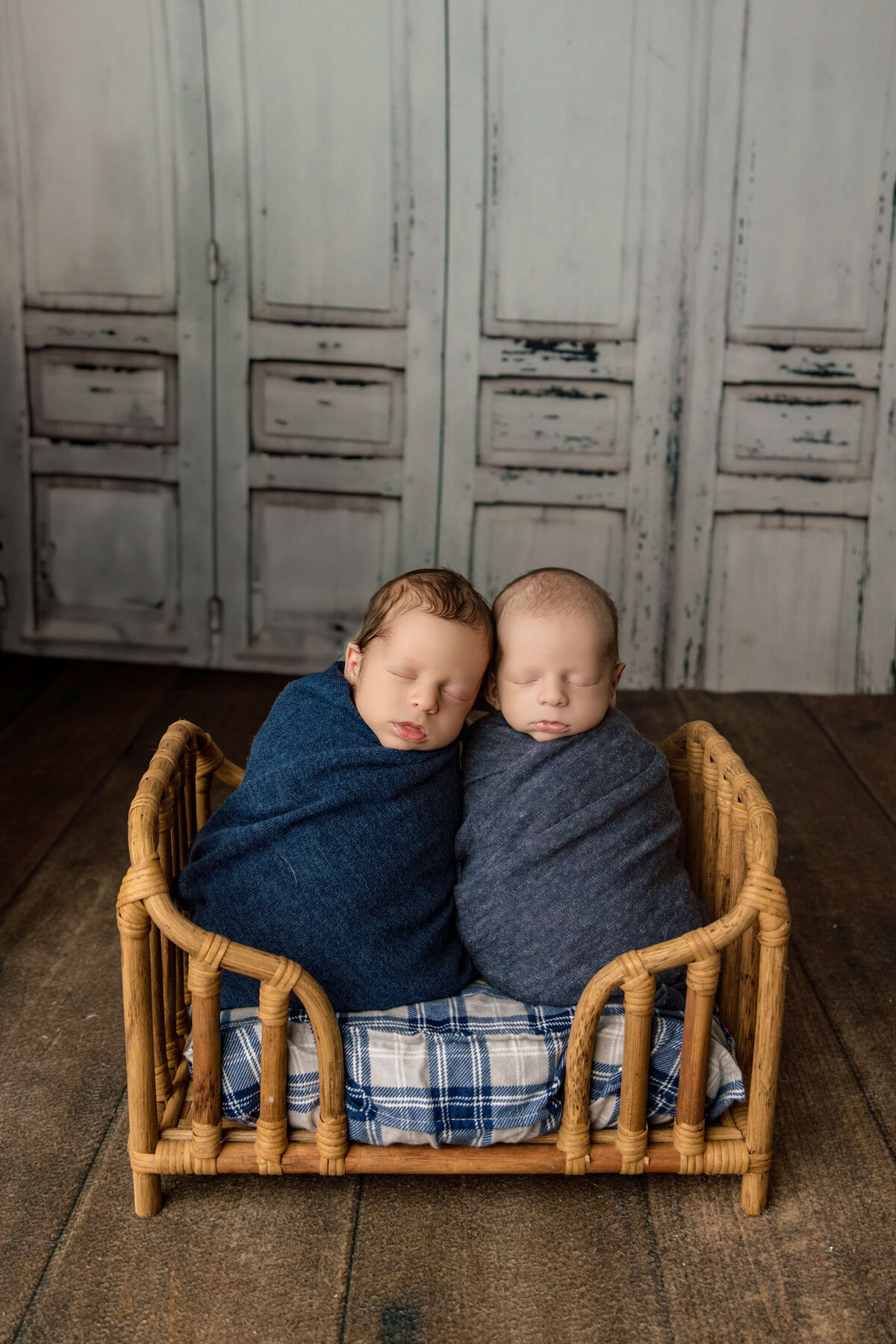 Two newborns peacefully sleeping side by side in a wicker crib. One baby is wrapped in a dark blue blanket, while the other is swaddled in gray. The crib sits on a plaid fabric, and a rustic wooden backdrop adds warmth to the scene.