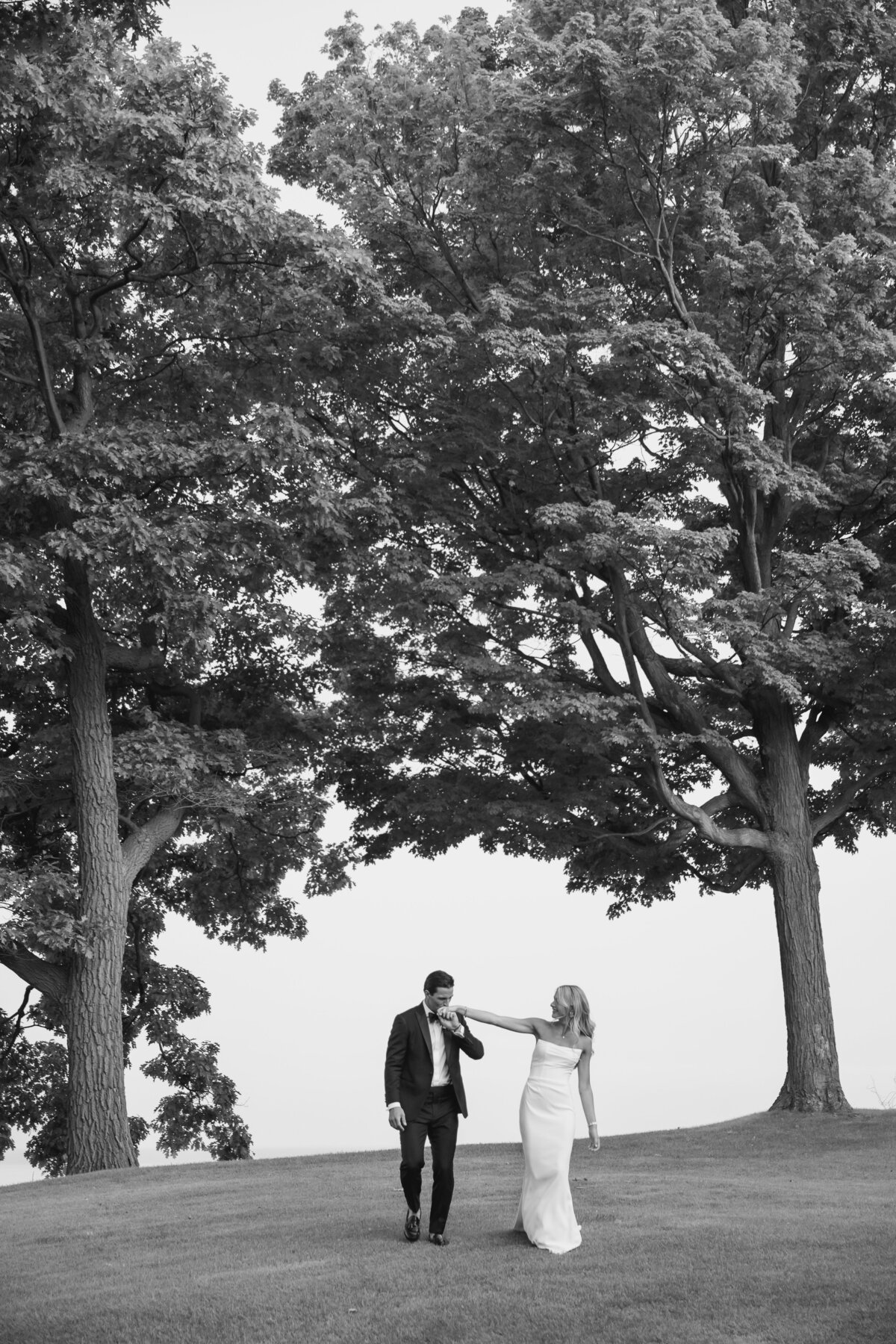 Black and white image of a couple on their wedding day walking down a hill. The groom is kissing the bride on her hand.