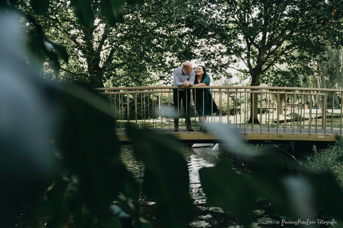 A candid, medium shot of a couple standing on a rustic, golden bridge over a small creek. The man, with a shaved head and a casual shirt, leans over the railing to look at the woman. She is wearing a dark green dress and smiling as she looks at him. They are framed by dense green trees and foliage, with dappled sunlight creating a relaxed, intimate atmosphere.