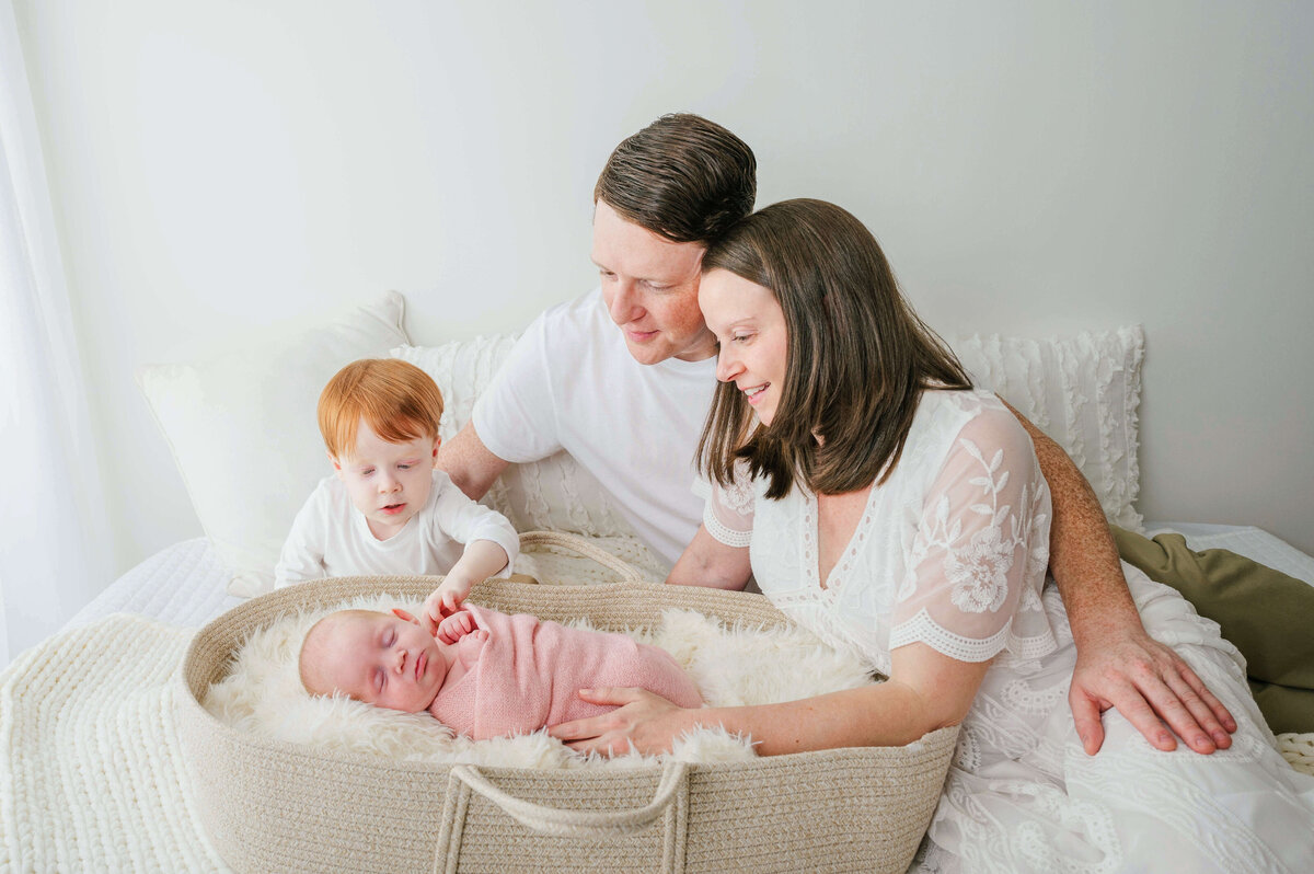 Brunette mom in a cream dress, and her husband and son sitting on a bed in front of a sheer curtain looking down at their newborn in a moses basket. 