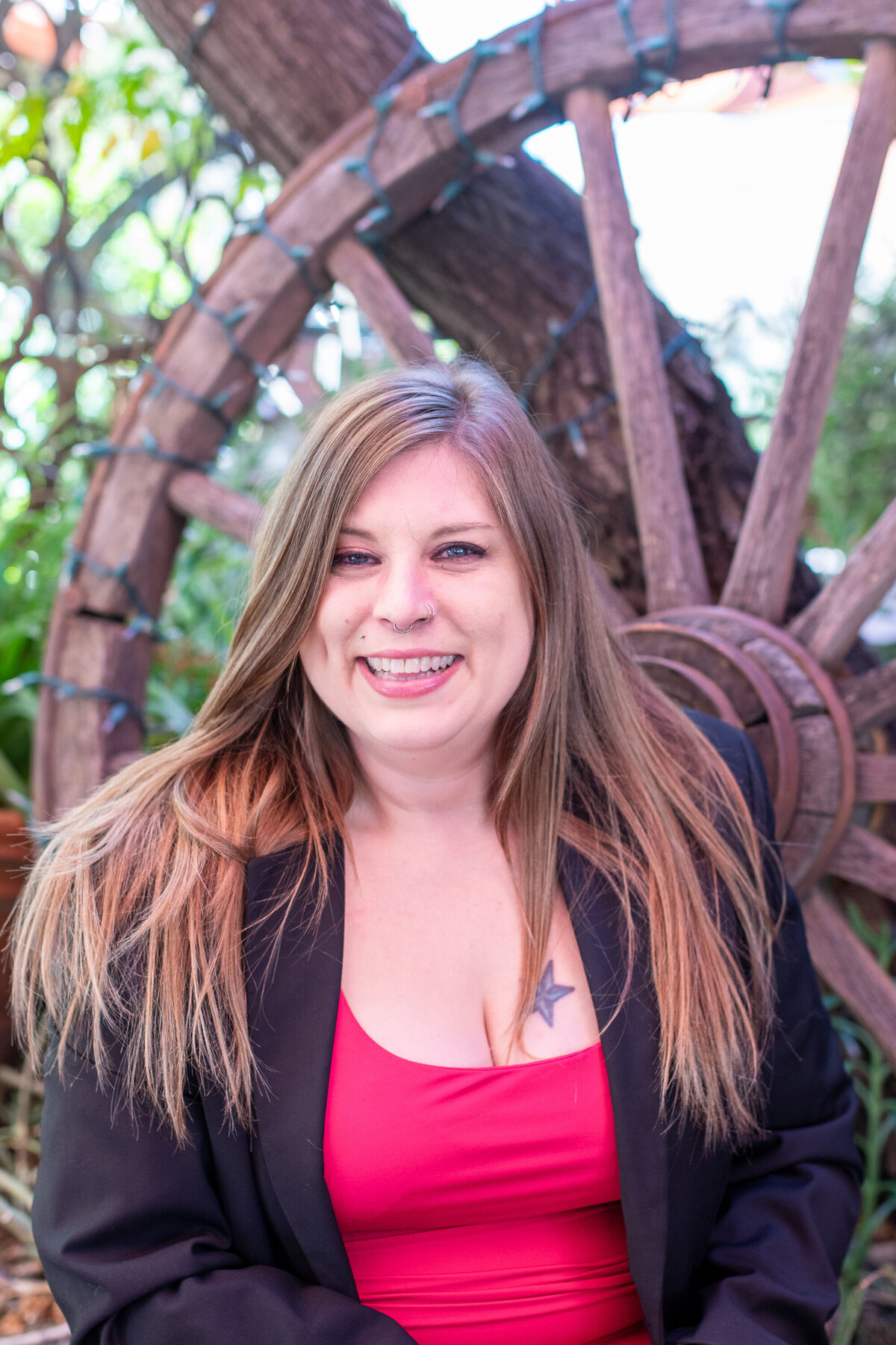Smiling woman with long hair wearing a black blazer and red top, posing outdoors in front of a rustic wooden wagon wheel, photographed by Vyrl Photo, showcasing Tucson brand photography.