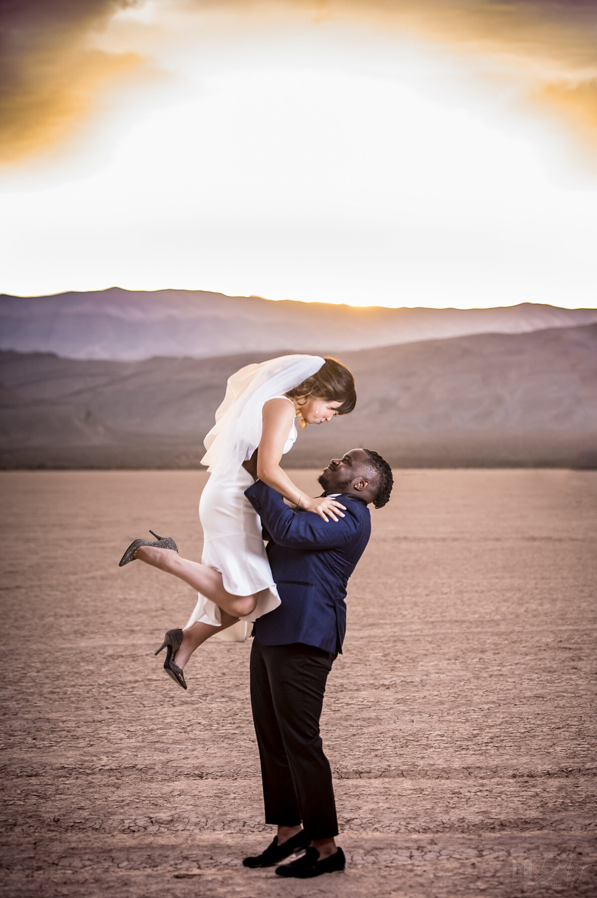 Groom lift his new bride up in the air las vegas elopement on the dry lake bed  at golden hour groom in blue suit jacket and black  pants  las vegas elopement eloping in vegas  las vegas wedding photographers las vegas wedding photography mk delacy photography