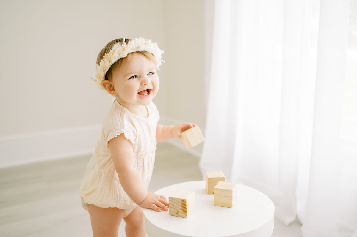 Baby girl stands at stool and plays with wooden toys during simple baby studio session in Raleigh photography studio