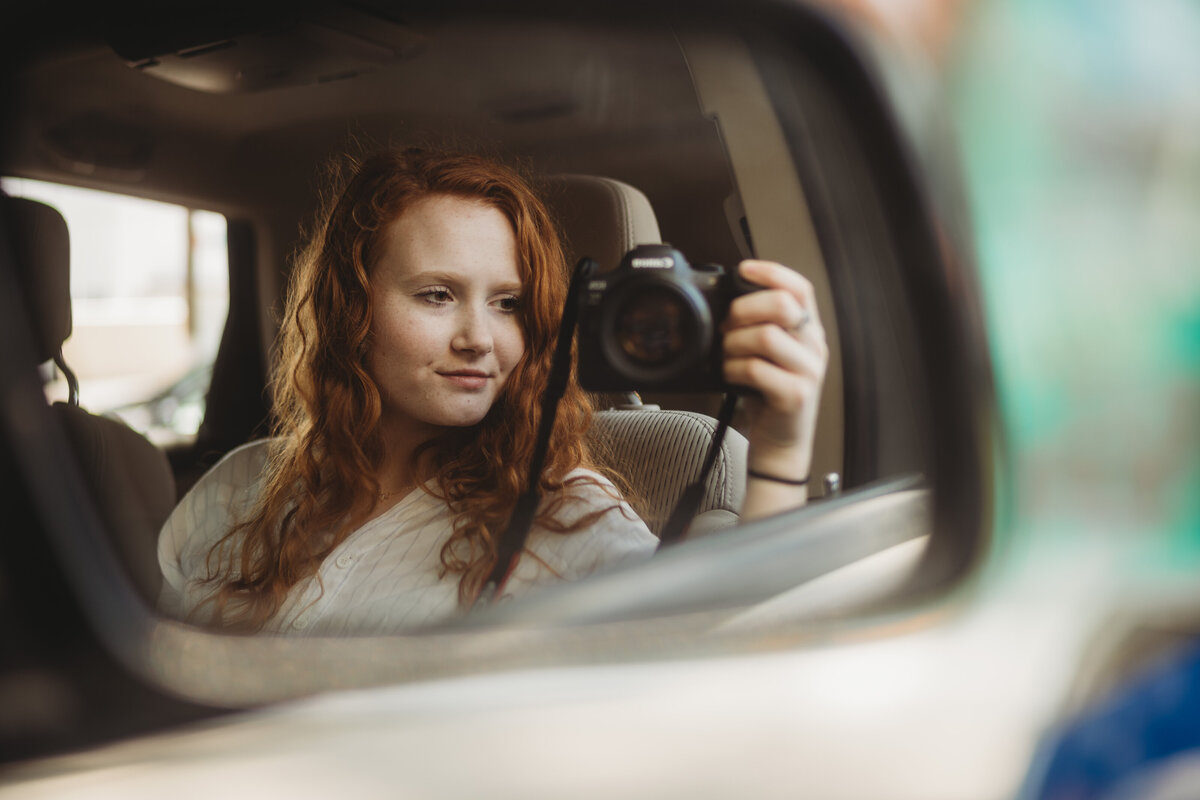 A red headed girl holds her camera out the window of a car and takes a photo of herself in the side mirror.