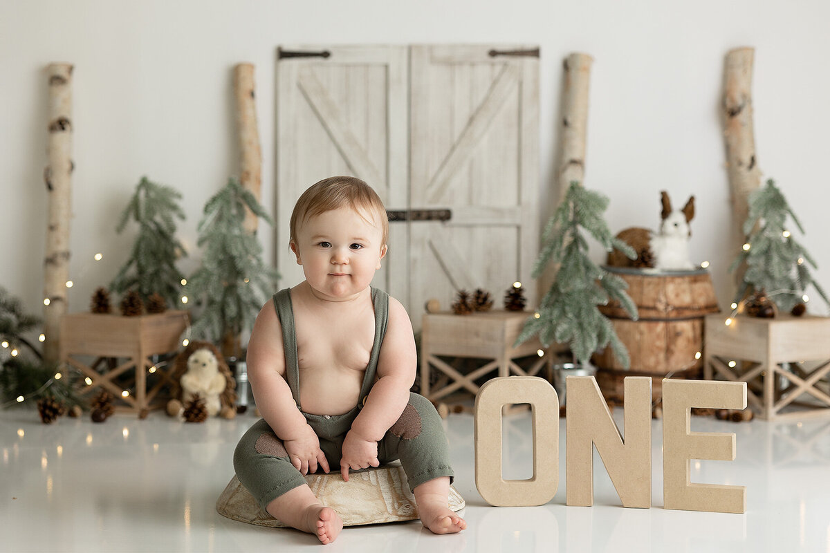 columbus-and-dayton-ohio-first-birthday-cake-smash-photographer-baby-boy-in-green-suspender-pants-with-simple-woodland-background-and-one-letters-amanda-estep-photography