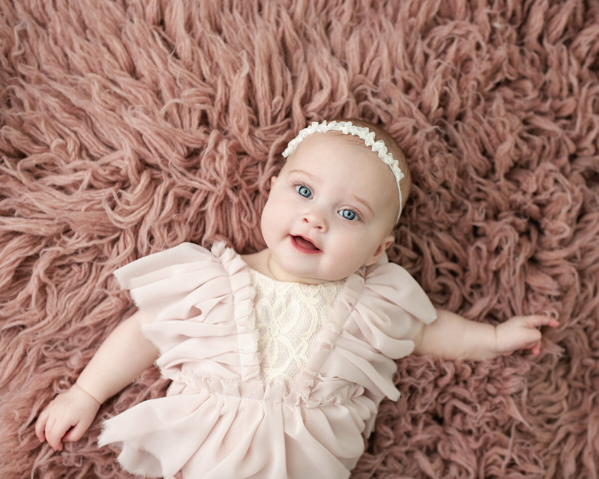 Smiling baby girl milestone portrait with soft pink textures and delicate ruffles—pure sweetness.